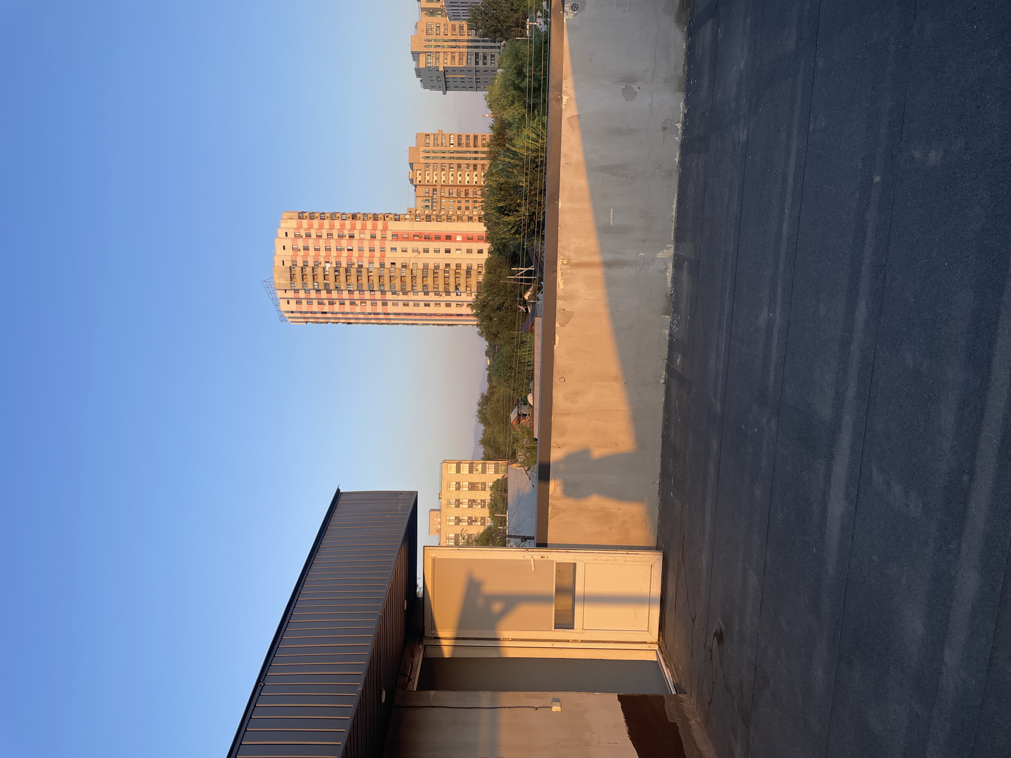 The image depicts a rooftop view overlooking a cityscape, likely in Almaty, Kazakhstan. The foreground features a dark, asphalt-like rooftop surface with visible seams and some lighter colored markings. To the left, a portion of a building with corrugated metal siding and a cream-colored wall with brown trim is visible. An open white door with a glass pane in the lower half casts a long shadow. A silhouette of a person is visible on the right side of the wall. In the background, several high-rise apartment buildings are present. The tallest building, with a pink and white checkered facade, stands out. Other buildings of varying heights and architectural styles are scattered amidst lush green trees. The sky is a clear, light blue, indicating it is daytime, possibly late afternoon due to the long shadows. The overall impression is a urban scene bathed in warm, natural light.