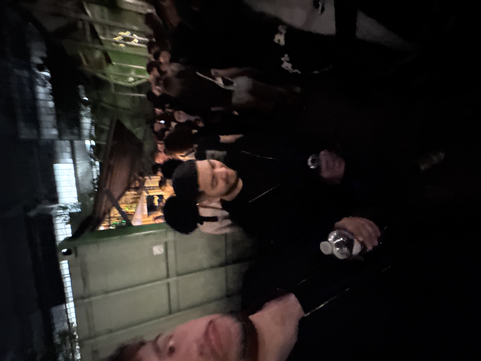 The image captures a night scene in an urban outdoor setting, likely in Paris, France. In the foreground, a person's face is partially visible on the left, indicating a close-up perspective. The central figure, dressed in dark attire, holds a plastic water bottle in their right hand. Other individuals are present in the blurry background, suggesting a gathering or crowd.
The setting includes a building facade with indistinct architectural details and a prominent large green industrial bin or dumpster to the left of the central figure. Artificial lighting is scarce, contributing to the overall dimness of the scene, typical of nighttime. The ambiance points towards a casual public event or social gathering in an urban environment. No discernible text is visible.