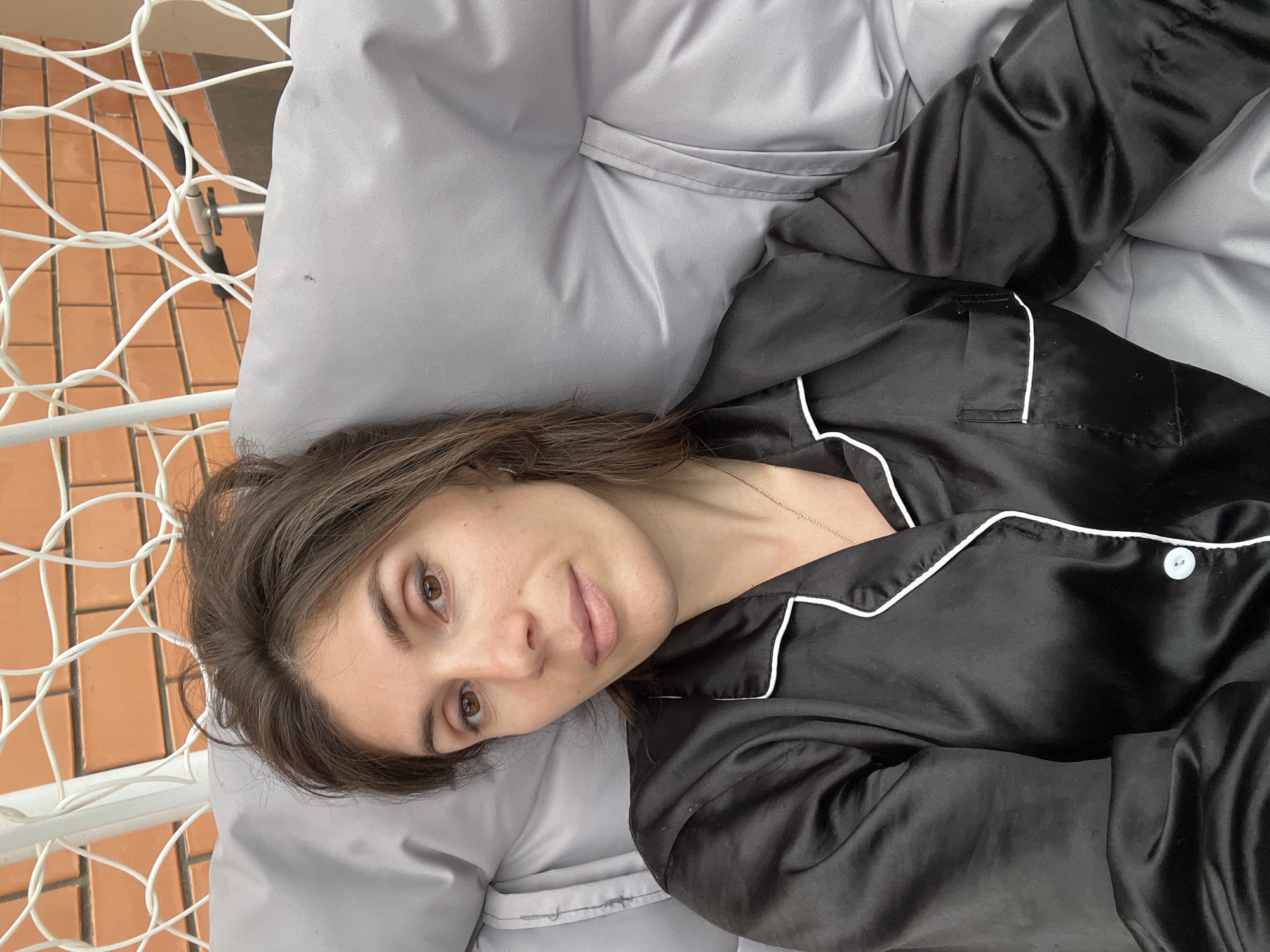 A medium close-up shot features an adult female, appearing to be in her late 20s to mid-30s, seated outdoors in the city of Kaspiysk, Russia. She has medium brown hair, casually styled and slightly disheveled, parted near the center. Her brown eyes are open and directed straight at the viewer, with subtle dark eyeliner on the upper lids. Her eyebrows are dark and well-defined. Her mouth is closed, forming a neutral to slightly pleasant expression, and her skin tone is fair.
She is wearing a black, satin-like long-sleeved pajama top with contrasting white piping along the collar, front placket, and sleeve cuffs. Two small white buttons are visible on the placket. A thin gold-colored chain with a small pendant hangs around her neck.
The woman is leaning back against a light gray, textured fabric cushion, which appears to be padded and is likely part of an outdoor furniture piece. Behind her head and to her left, a white, open-weave net-like structure is visible, consistent with a hanging swing chair or hammock. In the upper background, a reddish-orange brick wall is present, laid in a stretcher bond pattern, indicating an outdoor residential setting. To the right of her head, behind the cushion, a metallic fixture with a small black handle or grip is partially visible, likely part of the furniture or a nearby outdoor apparatus. The lighting suggests natural daylight.