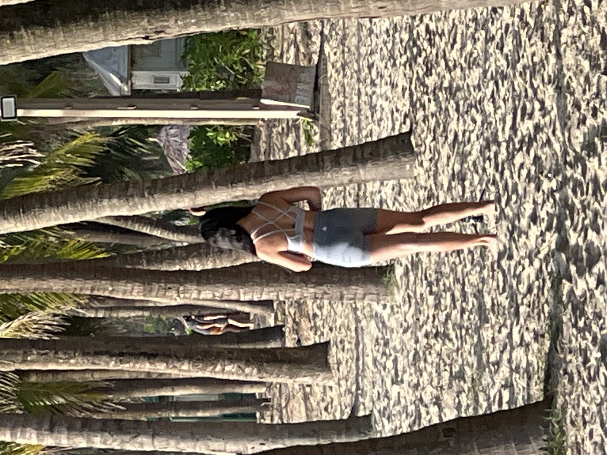 A woman is walking on a sandy beach lined with palm trees. She is wearing a grey sports bra and grey shorts. In the background, two other people are visible, and further back, a small building with a thatched roof can be seen. The scene appears to be in Đà Nẵng, Vietnam, known for its beaches and palm tree-lined coast.