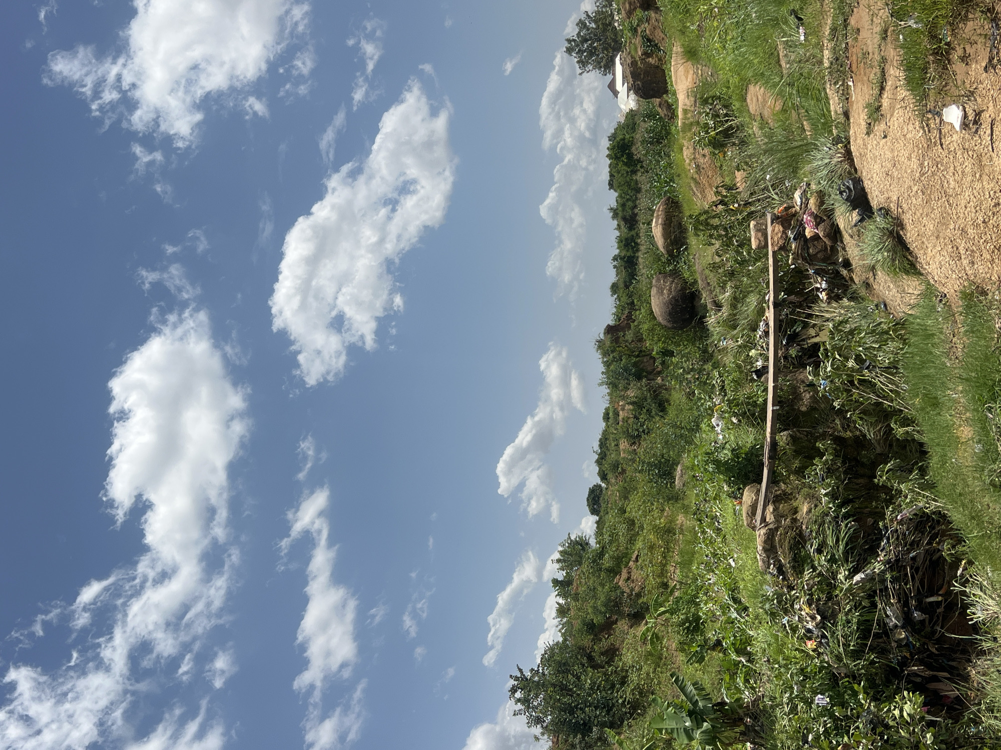 The image depicts an outdoor scene in Jos, Nigeria, characterized by a juxtaposition of natural elements and human impact. The sky is a vibrant blue, dotted with fluffy white clouds, suggesting a pleasant weather day. The landscape is hilly and covered in lush green vegetation, with some trees and what appear to be large, rounded boulders scattered across the terrain.
However, the scene is marred by the presence of trash and debris, particularly concentrated in the lower right portion of the image and along the edges of the grassy slopes. White plastic bags and other discarded items are visible amongst the plants and on the dirt path. In one area, a collection of items, including what looks like fabric or clothing, is piled near a wooden plank, further emphasizing the litter.
In the background, glimpses of buildings suggest a nearby settlement. The overall impression is of a natural landscape that is being encroached upon by waste, creating a somewhat discordant visual. The lighting and cloud formations suggest it is daytime, likely mid-morning or afternoon, under clear skies. No people are visible in the frame. There is no visible text in the image.