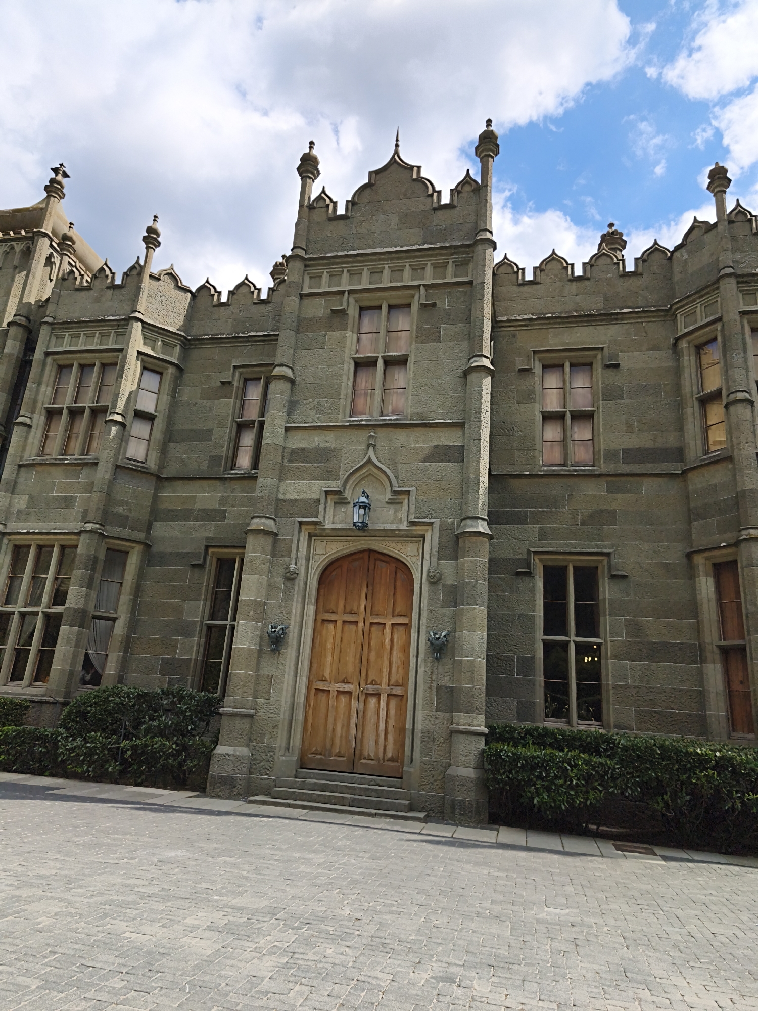 The image shows the Vorontsov Palace in Alupka, Ukraine. The palace is a large, grey stone building with a Gothic Revival style. It has a central tower with battlements and several smaller towers. The facade has many windows, and a wooden door is set in an arched opening with a stone lantern above it. Dark green bushes grow along the base of the building. The foreground is a paved courtyard of grey stone blocks. The sky is partly cloudy with blue patches visible.