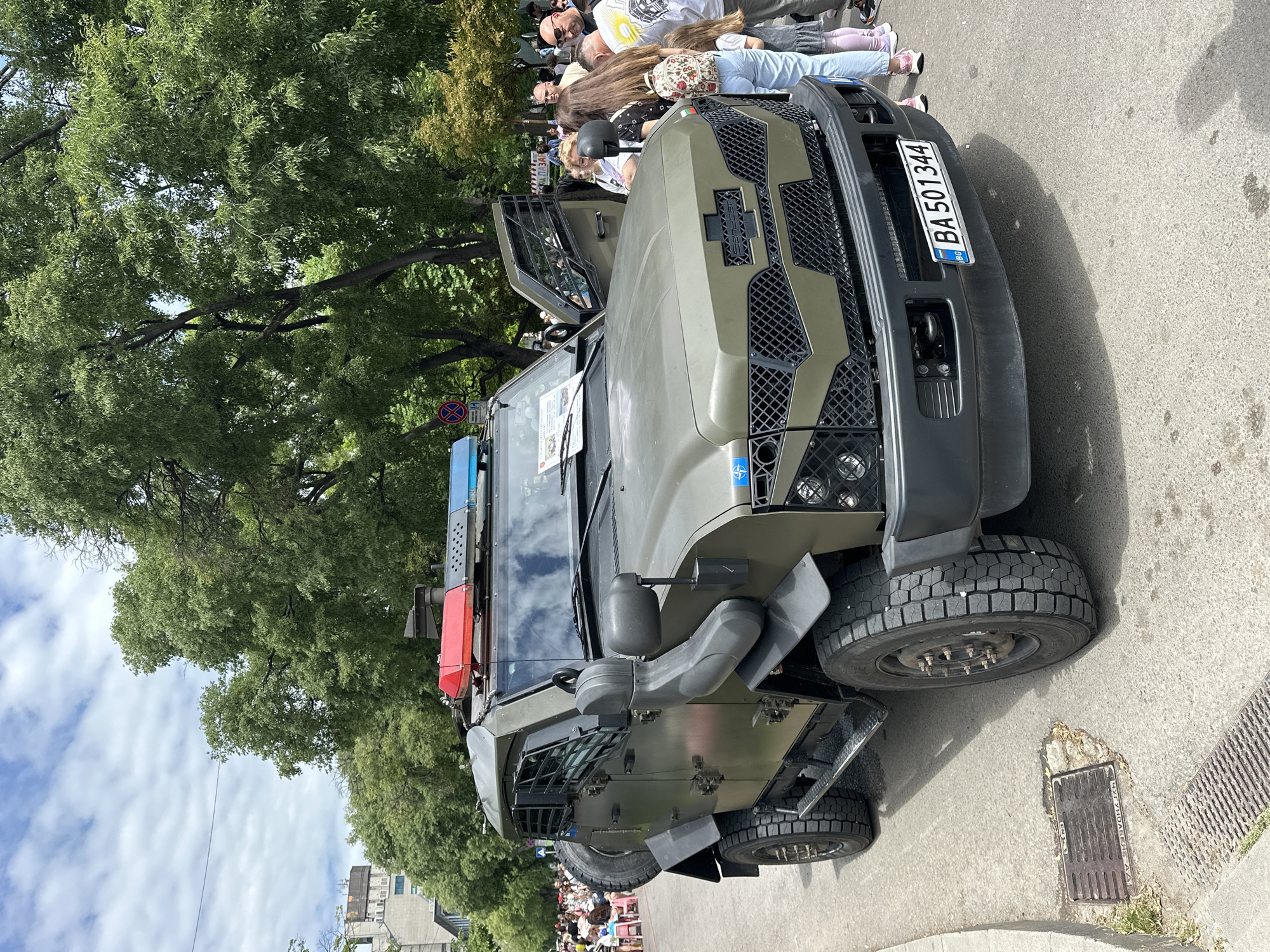 A close-up, medium shot shows a military vehicle parked on a paved road. The vehicle is dark green and heavily armored, with a grill over the front headlights. It has a Bulgarian license plate reading "BA 501344." A crowd of people stands on the sidewalk beside the vehicle, some looking at the vehicle and others at the camera. Behind the crowd are green trees. The sky is partly cloudy. This image likely depicts a static display of military hardware in a public place, given that the area features urban structures like curbs, sidewalks, and roads. There is no indication as to the location in Varna, Bulgaria.
