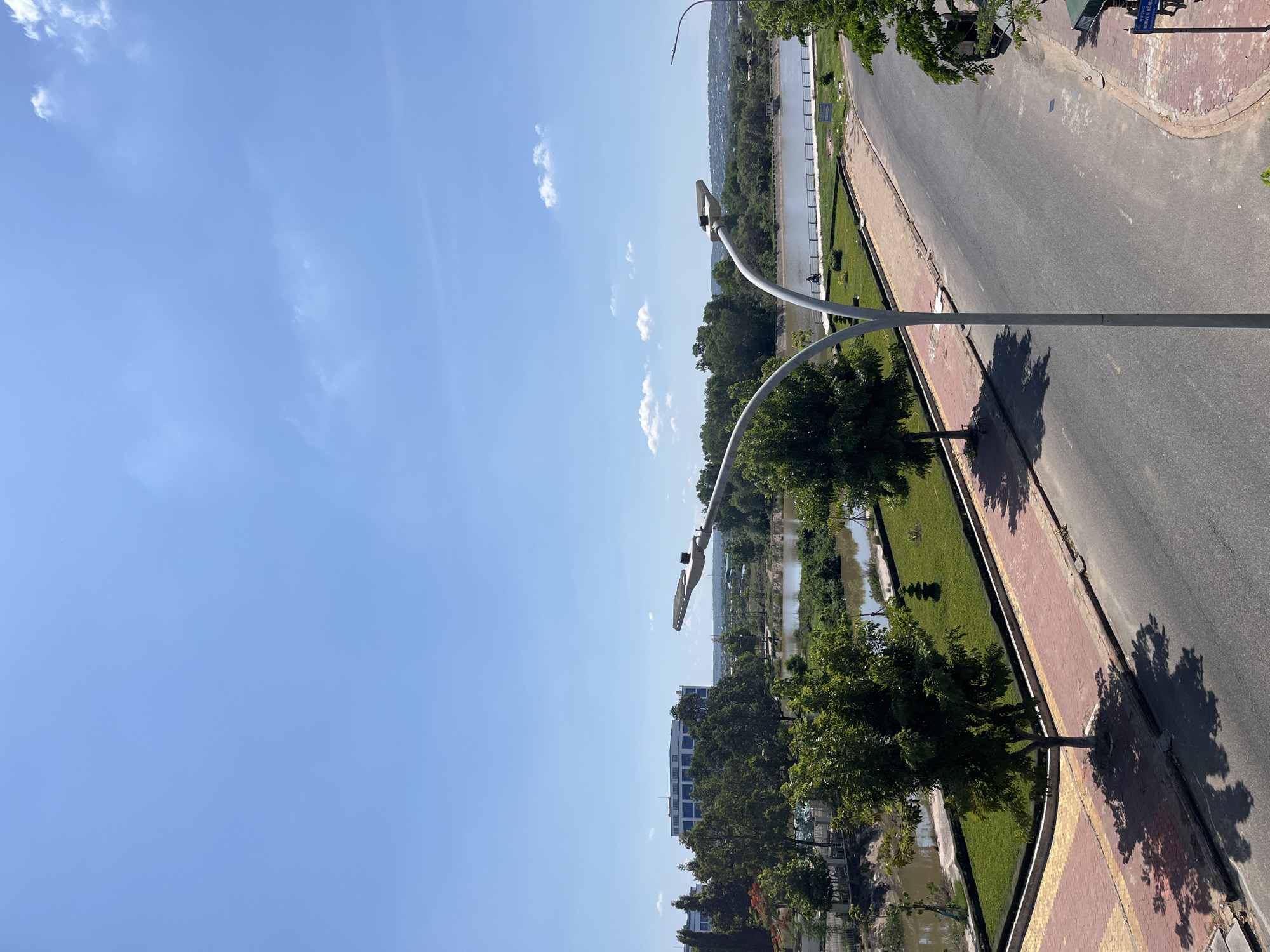 A bright, sunny day with a clear blue sky with sparse clouds is depicted in this image, likely taken from an elevated viewpoint looking out over a body of water and a city scene. In the foreground, a paved road curves to the right, with a sidewalk made of red and yellow bricks running alongside it. Two modern streetlights with curved, white poles are visible, one more prominently than the other.
Beyond the sidewalk and road is a green, grassy area bordered by a dark edging. A few trees with dense green foliage are scattered across this area, with one tree casting a distinct shadow on the sidewalk. In the middle ground, a calm body of brown water, possibly a river or lake, is visible, lined by a fence and more trees. In the background, a large, modern, multi-story building with a light-colored facade and numerous windows stands out against the greenery. Further in the distance, a hazy cityscape and rolling hills can be observed under the expansive sky.
A blue sign with white lettering is visible near the bottom right corner of the image, reading "PHƯỜNG HAM THẮNG NGHĨA". This indicates the location is in Ham Thang Ward, Vietnam. A single person on a bicycle is seen in the middle ground, riding along the path beside the water. There are no other prominent human figures or interactions in the scene. The overall environment appears to be a developed urban area bordering a natural water feature, with a focus on civic landscaping and modern architecture.