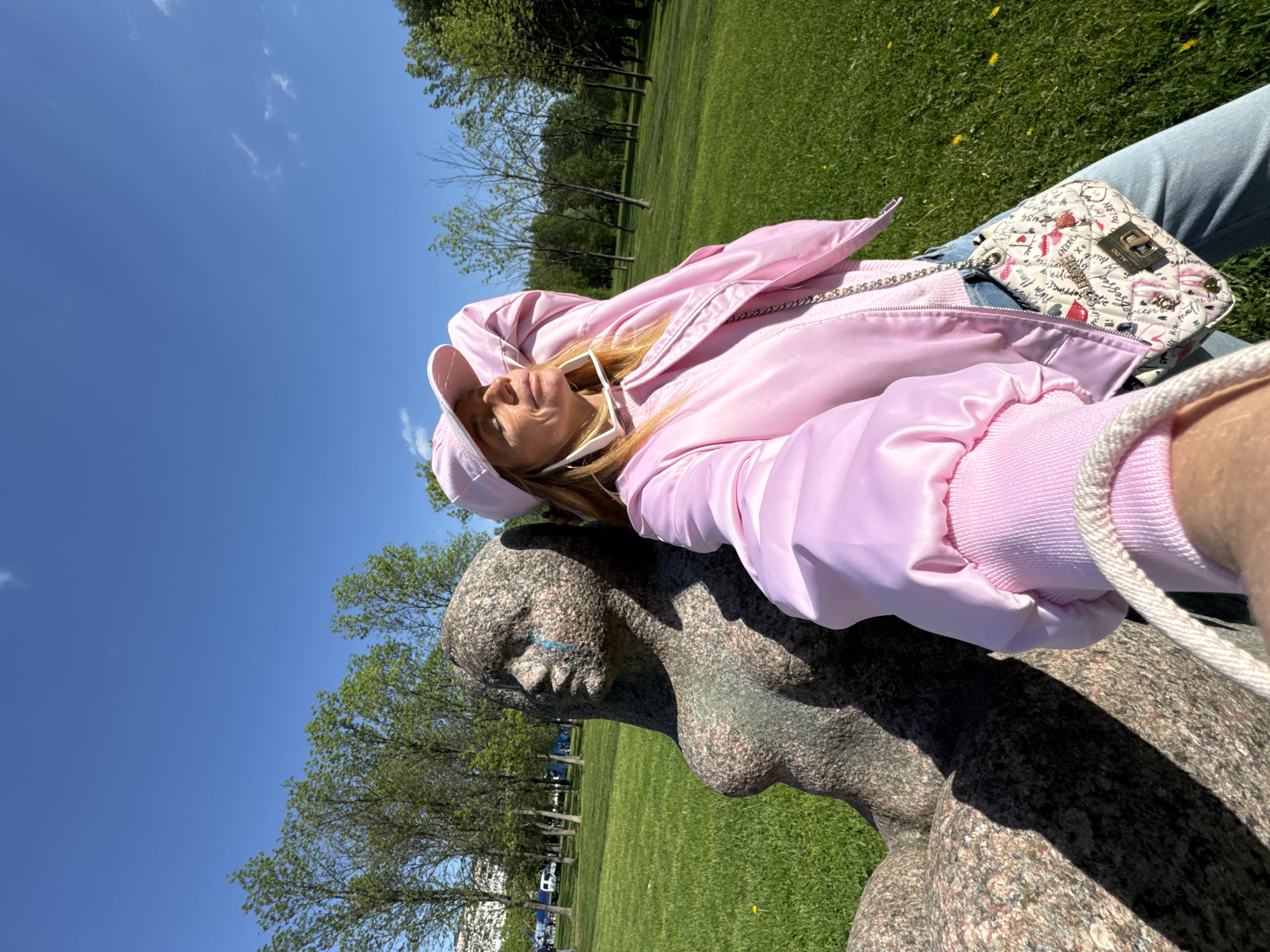 A woman in a pink jacket and hat is posing for a selfie with a granite sculpture of a female figure in a park. The statue features stylized facial features with blue accents. The background shows a green lawn, trees, a blue sky, and distant buildings that may be in Saint Petersburg, Russia. The woman is holding her phone at arm's length to take the photo. She also wears a purse with a white background and colorful accents.