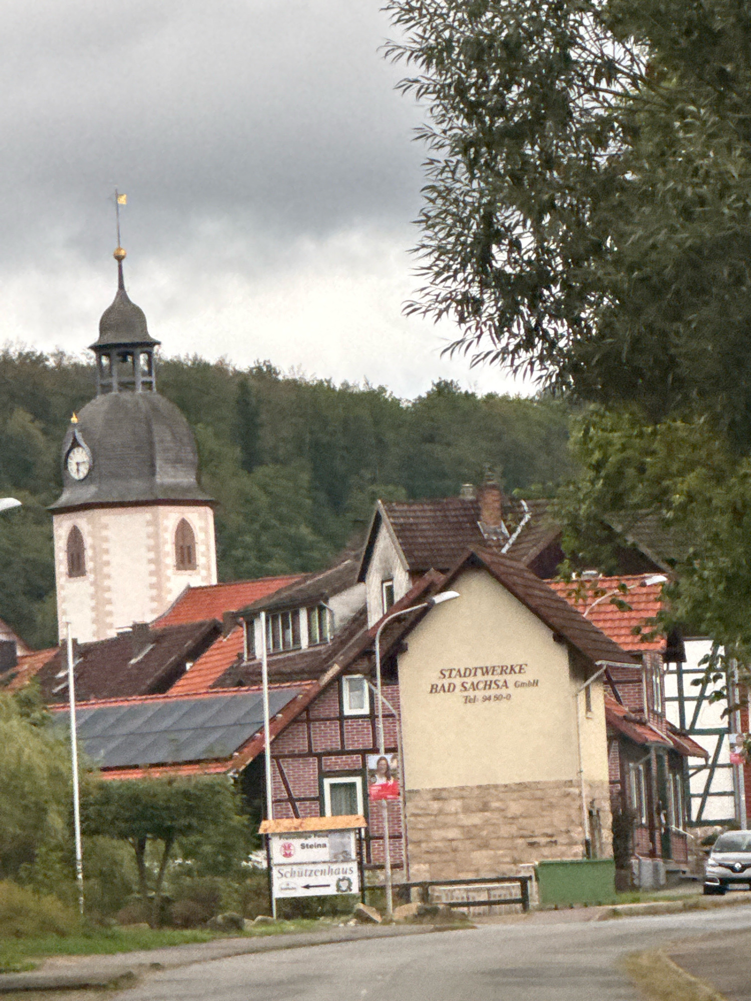 The image depicts a view of a town in Bad Sachsa, Germany, on an overcast day. In the foreground, a street is visible, with a grassy area and a sidewalk. Behind the sidewalk, there are several buildings. To the left, a tall, light-colored tower with a dark, domed roof and a golden finial stands out. A clock face is visible on the tower. Adjacent to the tower are other buildings with red-tiled roofs.
A building with a light-colored facade and a stone base has text that reads "STADTWERKE BAD SACHSA GmbH" and a phone number "Tel: 94 50-0". Another building features a black and white half-timbered design. Signs are visible, including one that says "Steina" and another with "Schützenhaus" and an arrow pointing left.
To the far right, a silver car is parked. Trees and dense green foliage form a backdrop to the town. The sky is heavily clouded, suggesting a cool or rainy day.