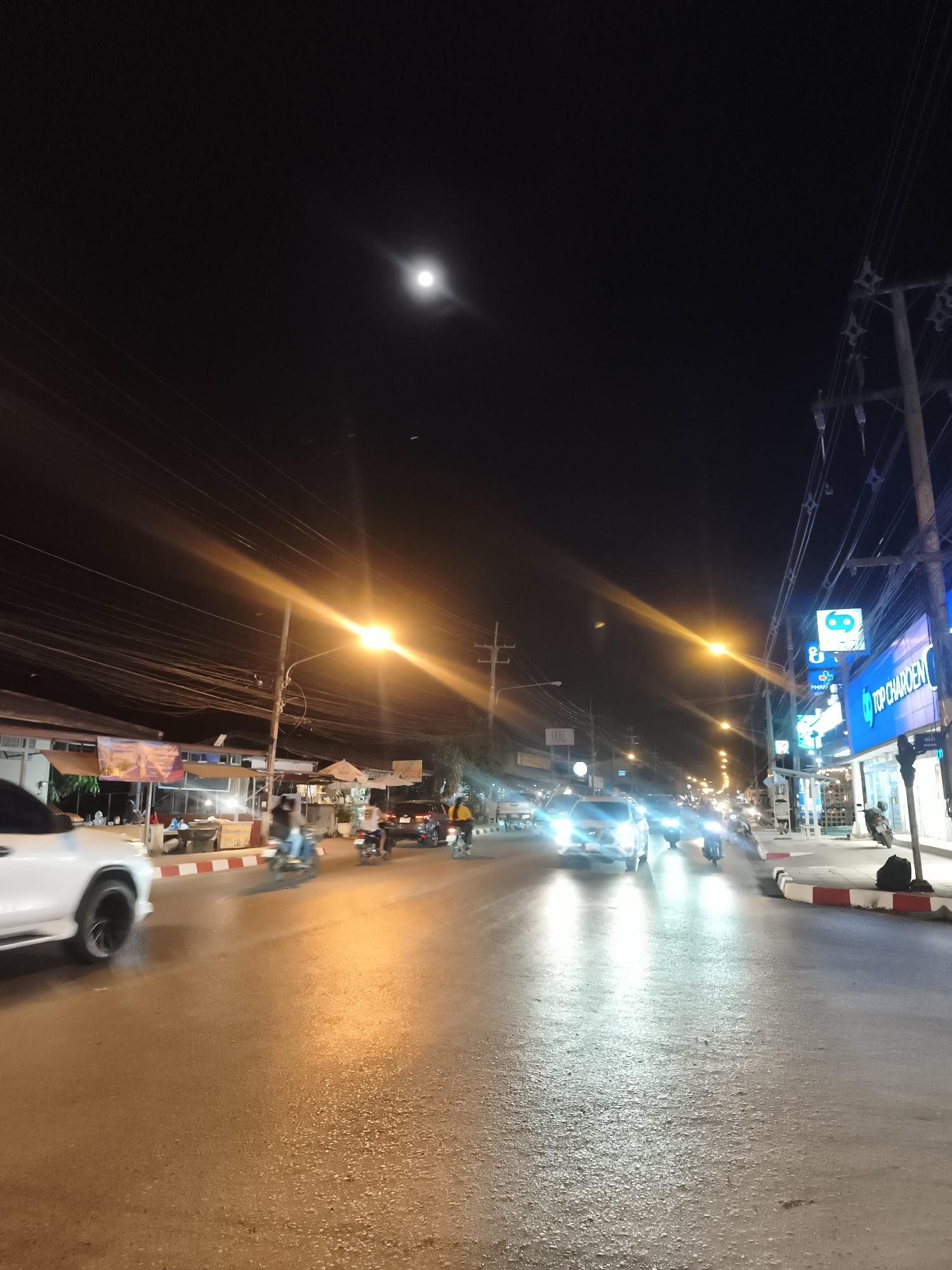 The image captures a vibrant night scene on a street in Baan Mae Nam, Thailand. The setting is an urban street illuminated by numerous artificial lights and a prominent moon overhead.
The dark night sky features a bright, full or nearly full moon centered in the upper frame, casting a soft glow. Below, the asphalt road appears reflective, likely from streetlights and vehicle headlights, possibly indicating a wet surface or just strong reflections. Red and white striped curbs line both sides of the street, separating the road from sidewalks and storefronts.
Traffic is visible, with several vehicles in motion. A white SUV is partially seen in the foreground left, its wheels blurred, suggesting movement. Further down the street, multiple cars have their headlights brightly lit, creating distinct light streaks and reflections on the road. Numerous motorbikes and scooters, some with riders, navigate the street or are parked along the side.
On the left side of the street, there are buildings with awnings, suggesting local businesses or eateries. On the right, commercial buildings are more clearly visible with brightly lit signs. A prominent blue and white sign for "TOP CHAROEN DENT" (a dental clinic) is visible, along with another partially visible "TOP CHAROEN" sign, likely an optical store. Other signs with Thai script are also present.
The scene is heavily illuminated by artificial light sources, including bright streetlights that create pronounced lens flare streaks across the image, vehicle headlights, and the glow from shop signs. A dense network of overhead power lines and communication cables crisscrosses the sky, attached to utility poles lining the street, a common sight in Thailand. Several people are visible, mostly as blurred figures on motorbikes or near the shops, indicating ongoing activity.