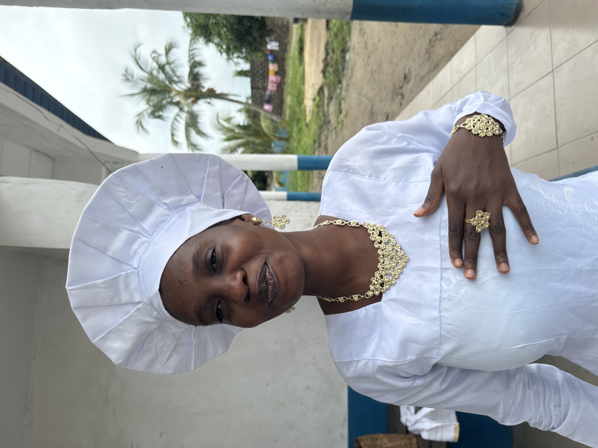 A woman in white religious attire stands outdoors in Badagry, Nigeria. She wears a white headdress, a white dress with a square neckline, gold ornate jewelry including a necklace, earrings, bracelet, and ring. Her right hand is placed over her chest. The background shows a cloudy sky, palm trees, a building with blue and white columns, and clothes hanging on a line.