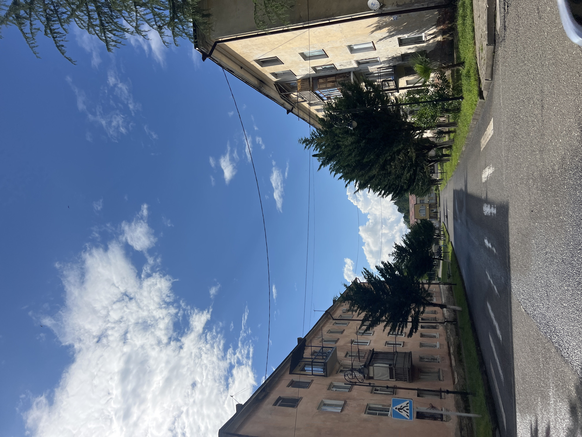 The image captures a view down a paved street lined with apartment buildings and mature trees. The sky above is a vibrant blue with scattered white clouds. On the left, a faded peach-colored apartment building has several windows and balconies. A pedestrian crossing sign is visible near the building. On the right, a light yellow apartment building also features windows and balconies, with a large evergreen tree in front of it and a palm tree further down. The street itself has white pedestrian crossing markings and appears to be wet in places, suggesting recent rain. The overall scene depicts a quiet residential street on a clear day.