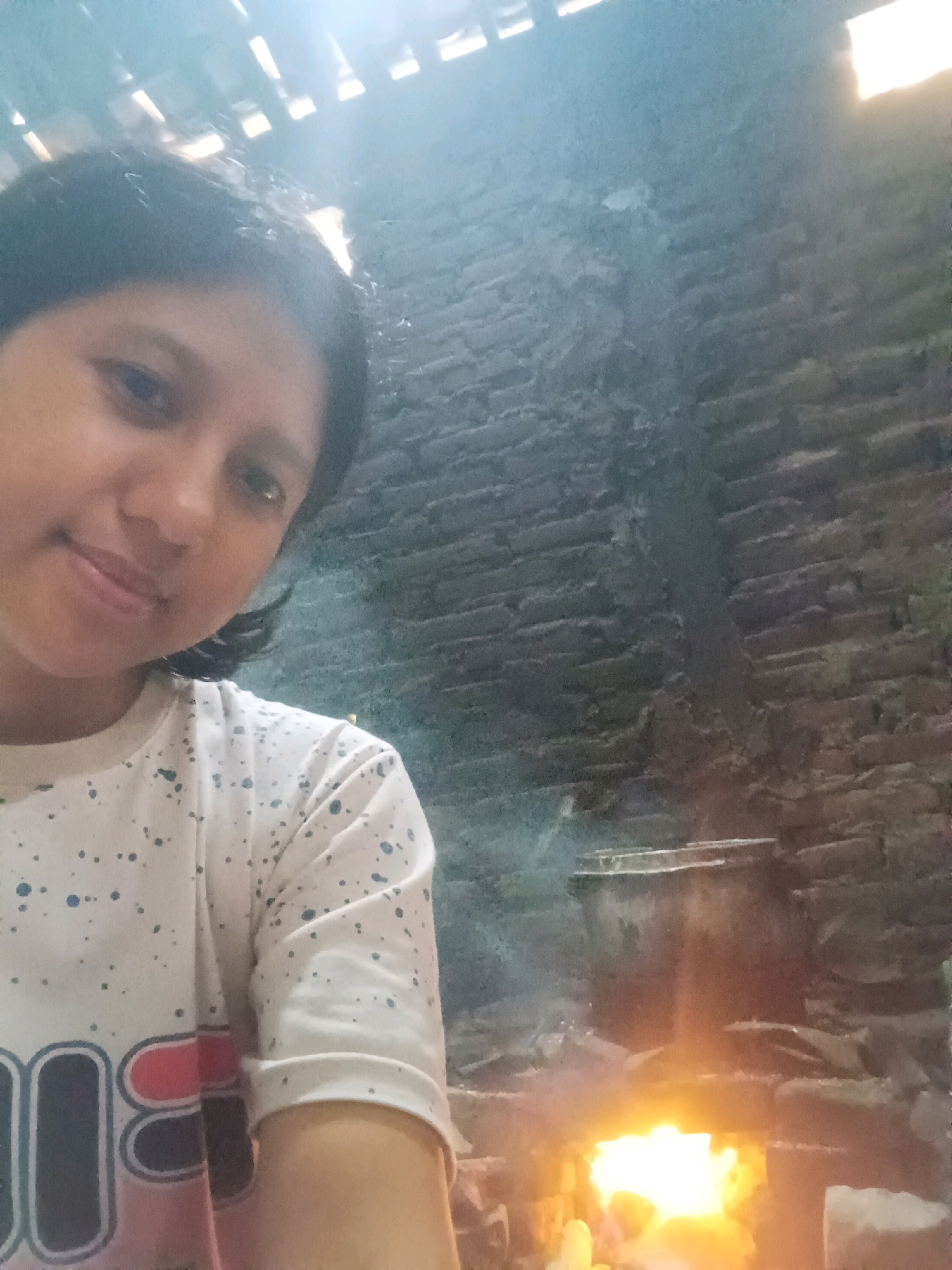 A person is seen in the foreground, taking a selfie. They are wearing a white t-shirt with a speckled pattern and a black and pink logo on the left side of their chest. Their hair is dark and shoulder-length. The background shows a rustic brick wall and a traditional stove with a fire burning, emitting smoke. The overall setting appears to be an outdoor or semi-outdoor cooking area, possibly in a rural location. The lighting suggests it is daytime, but the light source is not clearly identifiable, it could be natural light filtering in from above or artificial light from an unknown source. The image quality is moderate, with some blurriness and low resolution. The location is identified as CANGA'AN, Indonesia.