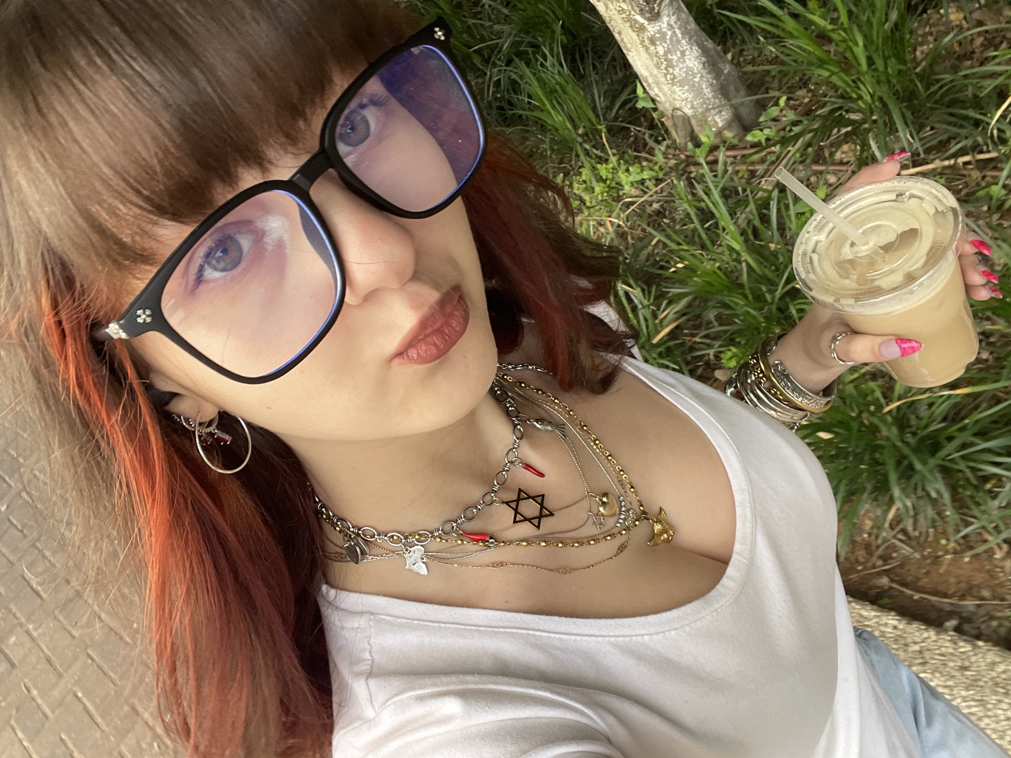 A woman takes a selfie outdoors. She wears oversized black-framed glasses with a light blue tint, a white top, multiple silver and gold necklaces, and bracelets. She holds a plastic cup with a straw, presumably filled with a cold beverage. The background includes a tree trunk and green vegetation. The setting appears to be a park or garden. The person is likely located in Raanana, Israel, based on the information provided.