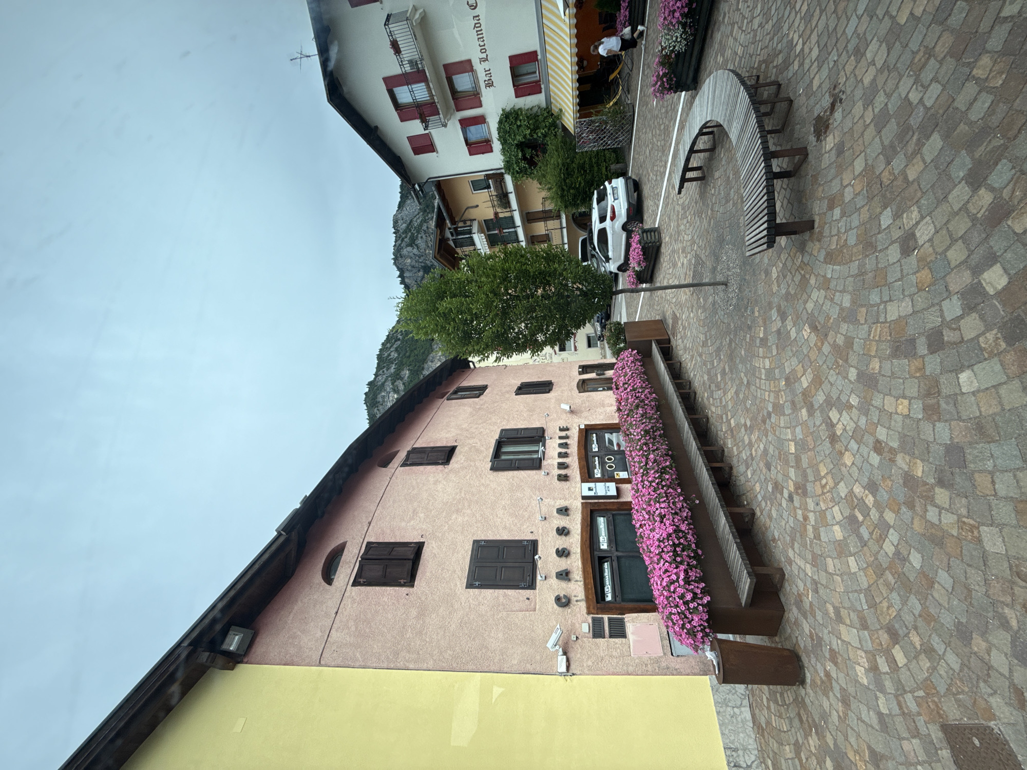 The image shows a view of a cobblestone plaza in Fai della Paganella, Italy. On the left, a pink building has the sign "CASSA RURALE" above its entrance. A long planter box filled with pink flowers runs along the front of the building, with a wooden bench in front of it. To the right of the pink building, a white SUV is parked. Further to the right, a building with a white facade and red shutters has a sign that reads "Bar Locanda Ca'". A person in a white shirt and black pants is walking near an awning with yellow and white stripes. In the background, mountains are visible under a cloudy sky.