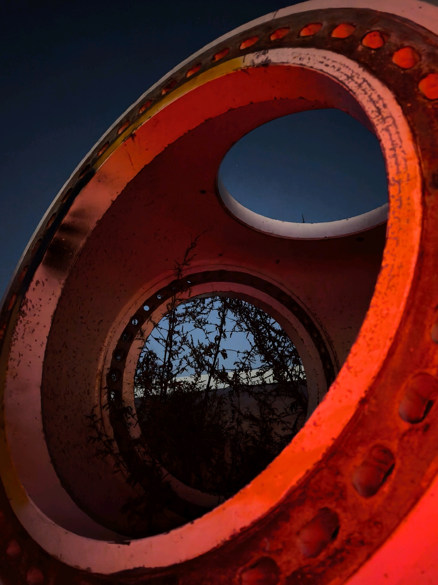 This is a close-up, low-angle shot of the interior of a large, circular, metallic object with a bright red outer coating. The object has two visible openings, one larger in the foreground and a smaller one further in the background, creating a tunnel-like effect. The red exterior is textured and appears aged, with visible signs of wear and possibly rust. Small circular indentations or holes are visible along the edges of these openings. Through the rear opening, a silhouette of dark, leafless branches and vegetation is visible against a twilight sky that transitions from a deep blue to a lighter hue near the horizon. The location is identified as Палыкавіцкі сельскі Савет, Belarus. The scene is devoid of any people or discernible activity, presenting a static, abstract composition focusing on the interplay of light, form, and texture within the constructed object and the natural elements beyond.