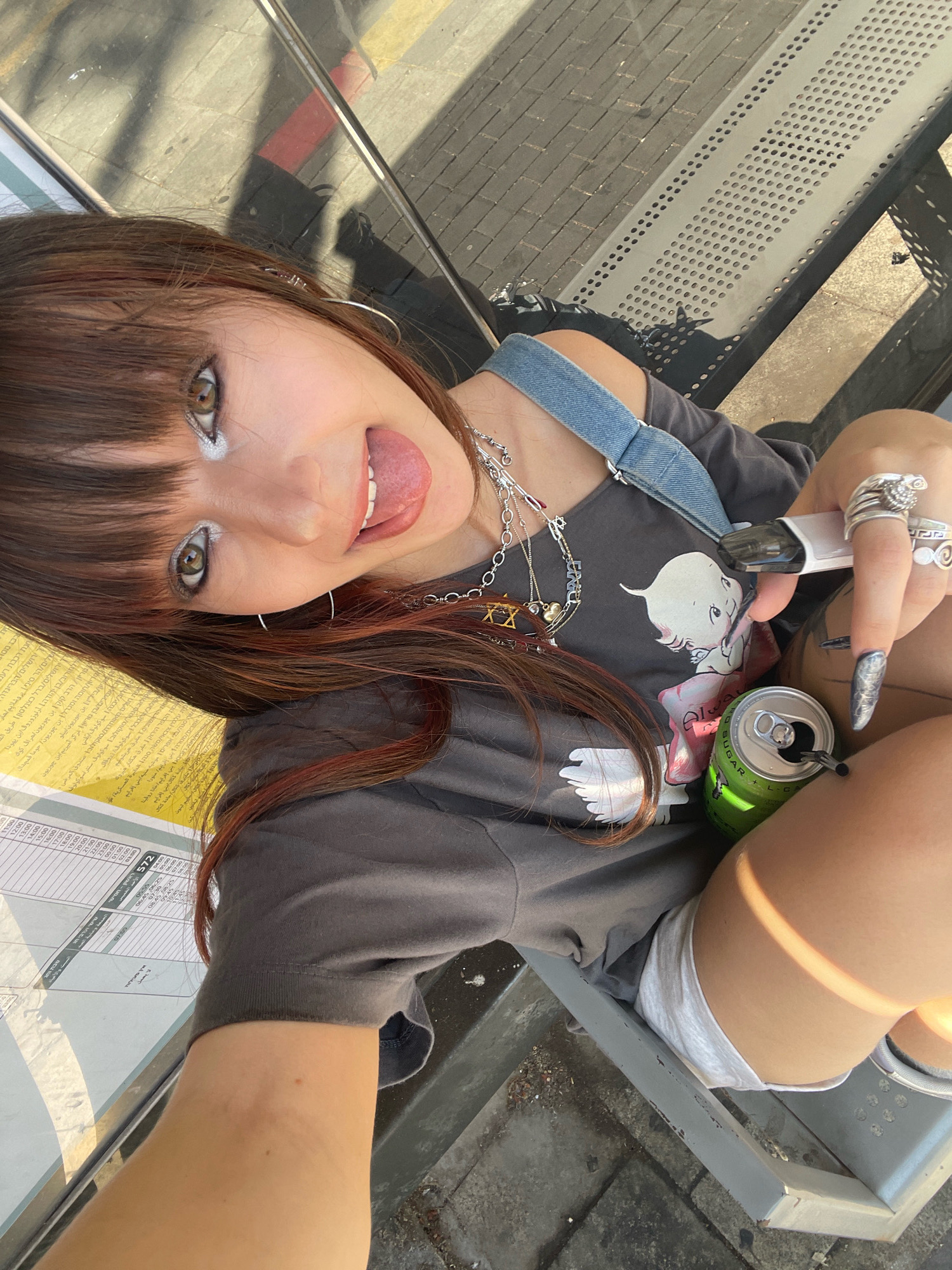 The image shows a young woman with brown hair and bangs sitting on a bench at a bus stop in Raanana, Israel. She is wearing a gray t-shirt with a cartoon graphic, white shorts, and a denim strap. She has on multiple rings and necklaces. She is holding a vape pen in her right hand and a can of green soda in her left hand. Her tongue is sticking out. Behind her is a bus schedule. The bench appears to be made of metal with perforated seating. The ground is paved with bricks. Sunlight casts shadows on the ground and the bench.