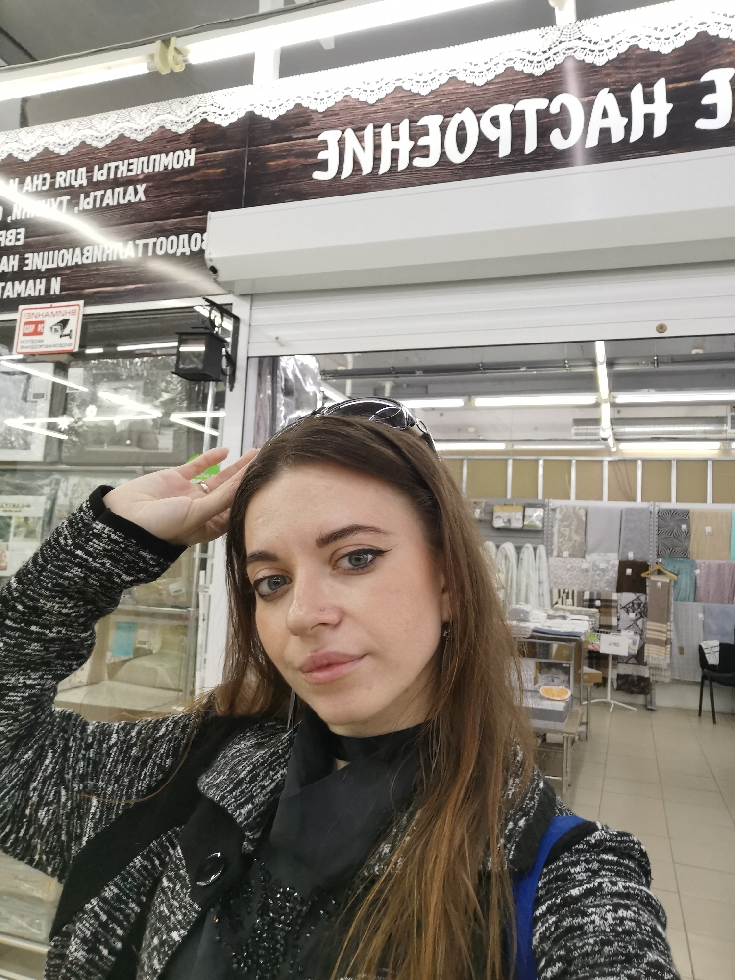 A medium shot captures a woman in front of a store. The background includes the store's display of home textiles and a security sign is posted. The woman is wearing a black and white patterned coat with a blue backpack strap visible. She has sunglasses perched on her head and is touching her forehead with her left hand. She appears to be inside the store. The location is Unknown, Unknown.