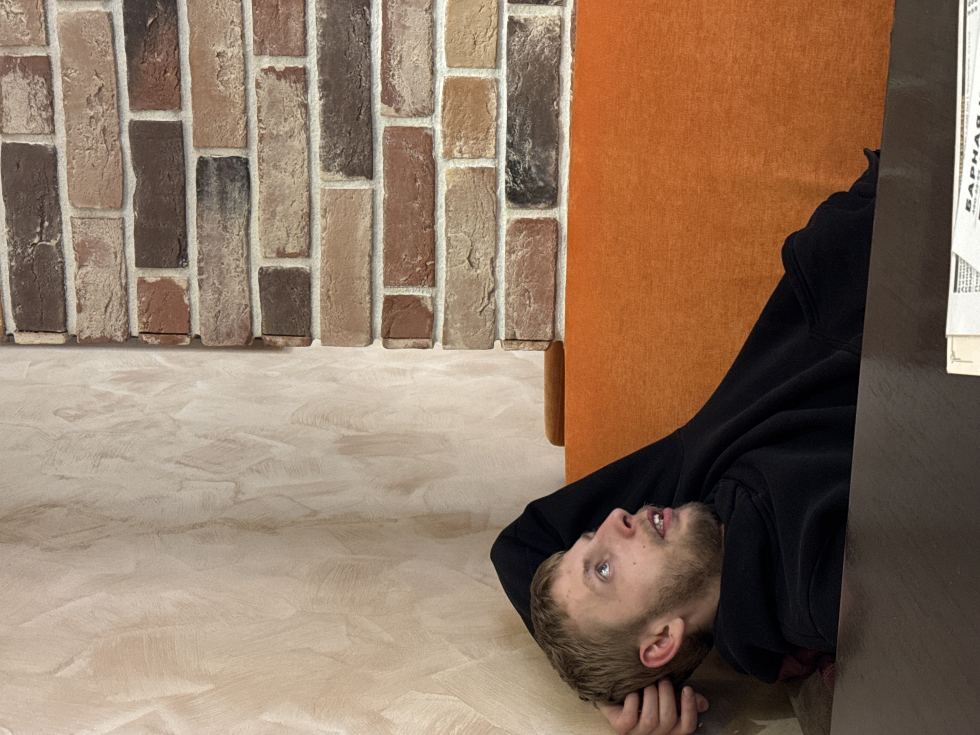 A man lies on his back on a light beige floor with a textured surface, wearing a black hoodie and resting his head on his hand. Adjacent to him are a dark brown table, an orange upholstered chair, and a textured brick wall featuring various shades of brown. A piece of paper with Russian text on it is on the table. The location is inferred to be Saint Petersburg, Russia, based on the Russian text present.