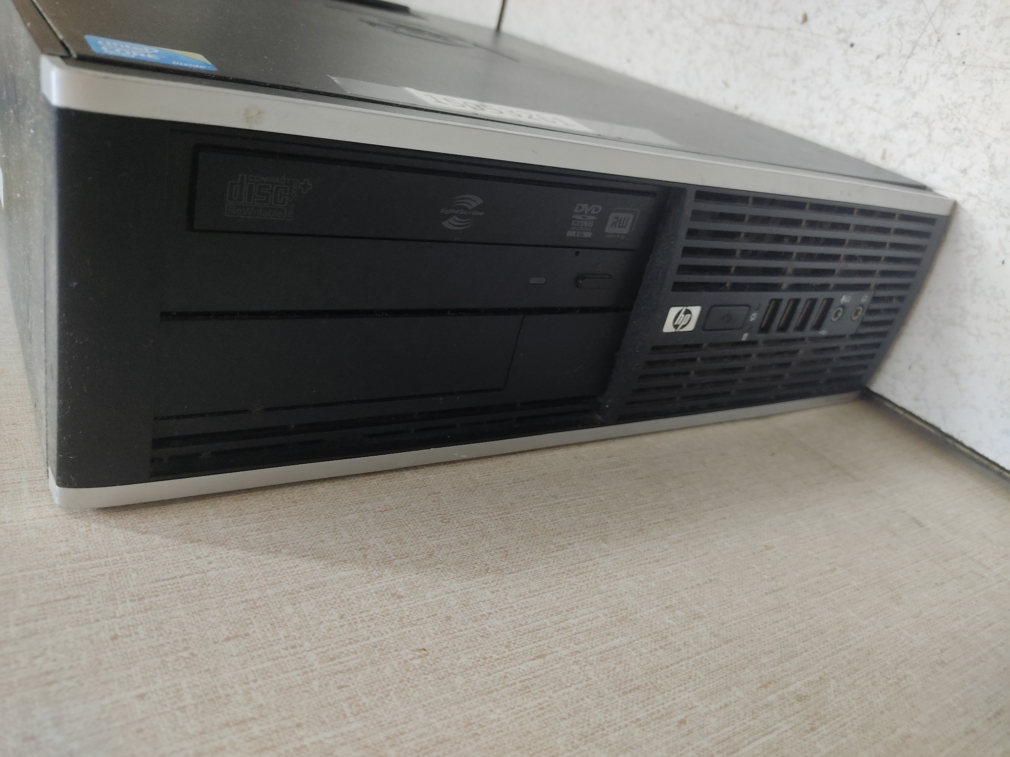 A desktop computer tower is positioned on its side against a lightly textured, beige wall. The computer is black with a silver trim around the front panel. The front panel features a DVD drive with a "disc+" label, a power button with an HP logo, and several USB ports. The scene is indoors, with the computer resting on a light-colored, speckled surface.