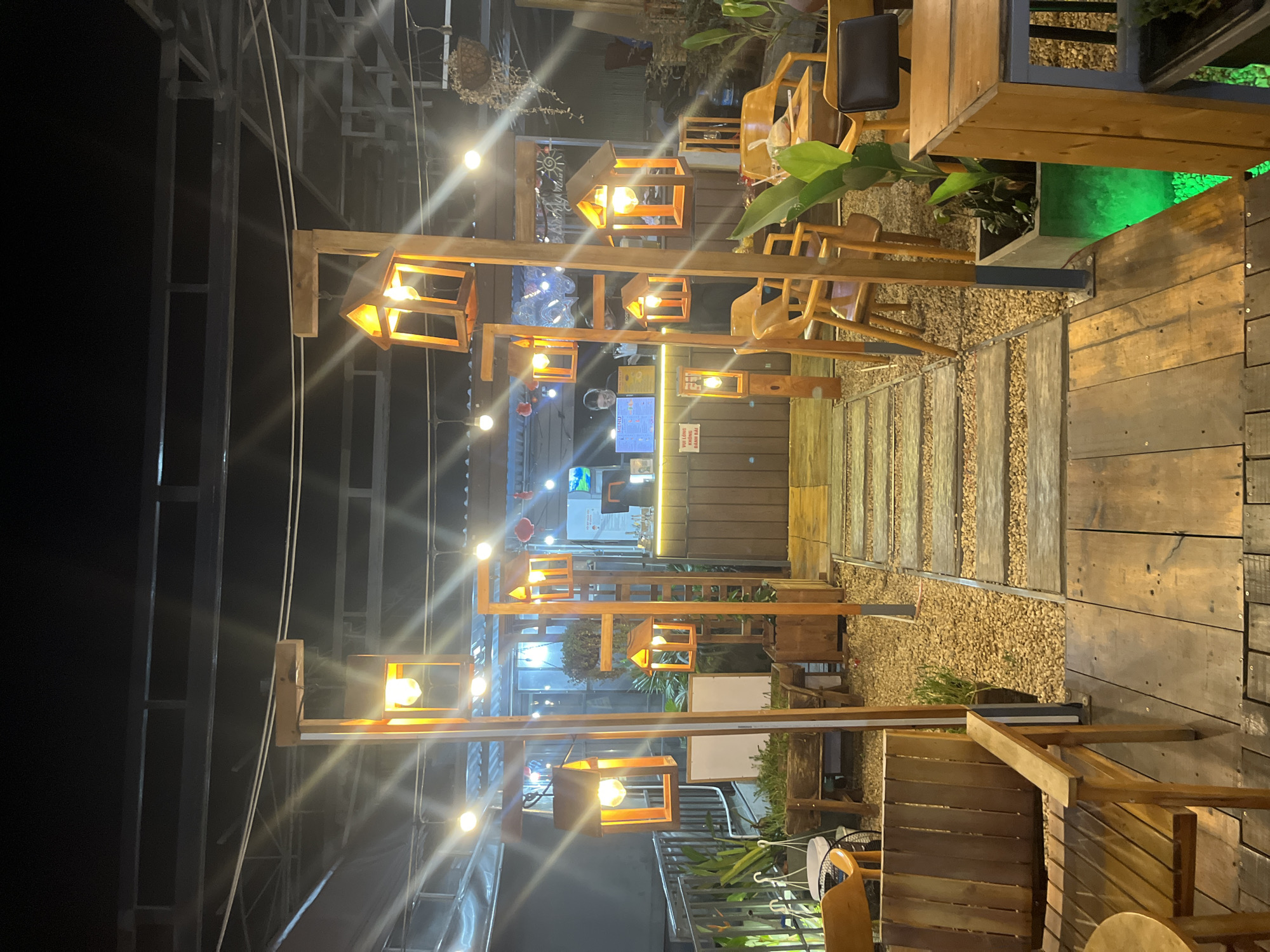 This image shows an outdoor seating area of a cafe at night in Da Nang, Vietnam. The scene is illuminated by warm, ambient light from numerous wooden lantern-style fixtures hanging from a metal frame structure. The cafe features a rustic, natural aesthetic with wooden furniture, including chairs and tables, scattered throughout the space. A pathway made of wooden planks interspersed with gravel leads through the seating area. In the background, a covered section of the cafe is visible with more seating and a counter area, where a person can be seen behind the counter. Plants are strategically placed around the seating, adding a touch of greenery. The overall impression is that of a cozy and inviting atmosphere, perfect for a relaxing evening. The time of day is clearly nighttime, indicated by the dark sky and artificial lighting. There is no discernible weather information. Some text is visible on a sign in the background, but it is too blurry to read.