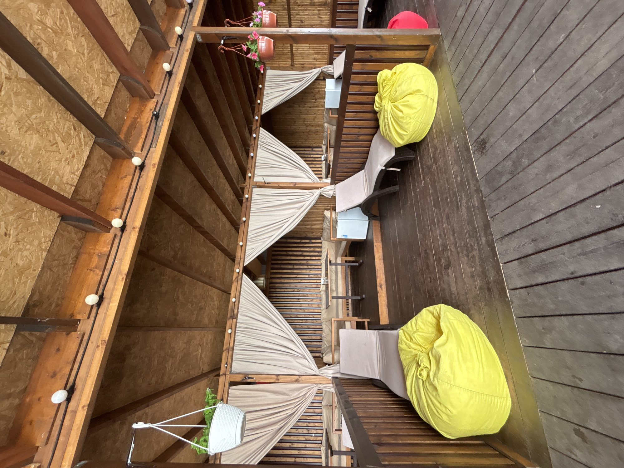 This media file captures an overhead view of a wooden outdoor seating area, possibly a balcony or a covered patio. The scene is oriented such that the wooden planks of the floor run diagonally from the lower left to the upper right.
The primary subjects are several pieces of outdoor furniture. Two light-colored lounge chairs are visible, one with a yellow beanbag resting on it, and another with a yellow beanbag beside it. In the background, a red beanbag is also present on a wooden surface.
The setting is a covered structure with wooden beams and what appears to be plywood or particleboard on the left side. Draped beige fabric partitions create a segmented, intimate feel to the space. String lights with small, round bulbs are strung along the wooden beams on the left. Hanging planters with pink flowers are visible at the top of the structure.
The time of day is not explicitly indicated, but the lighting suggests it is daytime. The weather appears to be mild, conducive to outdoor seating. There are no people visible in the frame. No text is discernable in the image. The location context provided is Sochi, Russia.