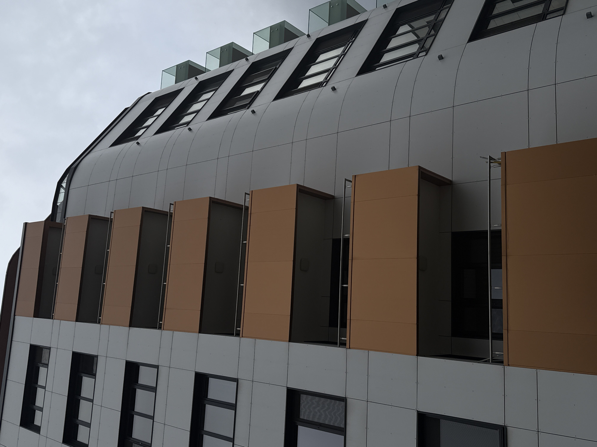 The image depicts a modern building in Sofia, Bulgaria, characterized by its unique architectural design. The building features a curved facade covered with gray panels, punctuated by rows of windows. The uppermost row consists of slanted windows topped with glass structures. A mid-level row showcases box-like structures with orange-brown panels, some of which appear to be open. The lower level exhibits rectangular windows with black frames set against a gray paneled wall. The sky is overcast.