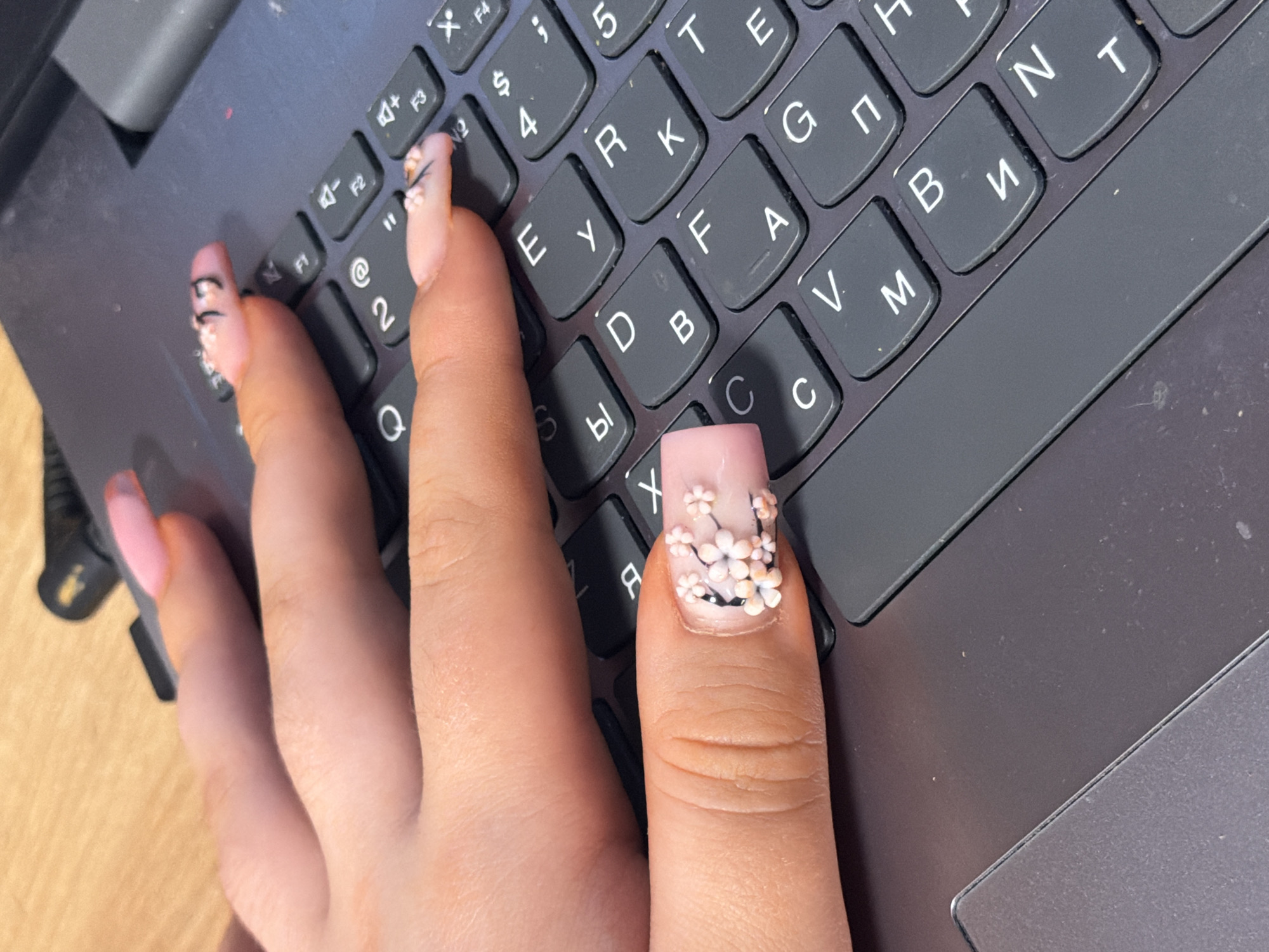 A close-up, overhead shot shows a person's hand with decorated fingernails resting on a dark grey laptop keyboard. The fingernails are long and painted with a light pink polish. The index finger has a detailed 3D floral design of small white and pink flowers with black stems. The middle finger has black lines and a pink design. The hand is positioned across several keys, including what appear to be "F1," "F2," "2," "@," "E," "Y," "D," "B," "R," "K," "A," "F," and "C." The keyboard is part of a laptop that is open. The background is a light-colored wooden surface. The scene does not provide any information about the city of Perm, Russia.