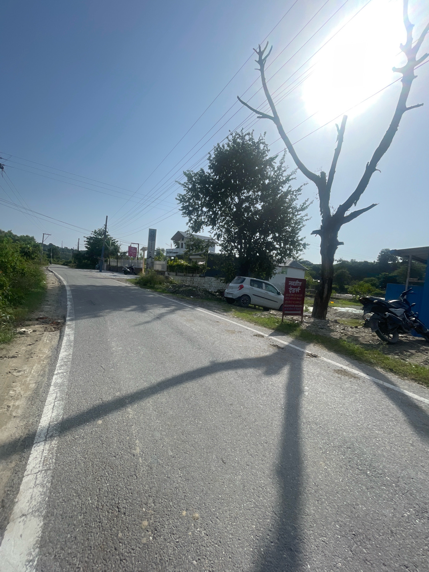 The image shows a view from a paved road in Kolhupani, India, on a bright, sunny day. The road curves to the left, with a white line marking the edge. On the right side of the road, a dark-colored motorcycle is parked. Further down the road, a white car is parked at an angle on the grassy shoulder. A red sign with text in an Indian script is placed near the car and a leafless tree. In the background, there are buildings and lush green trees. Power lines are visible stretching across the clear blue sky, and the sun is shining brightly, casting long shadows on the road.