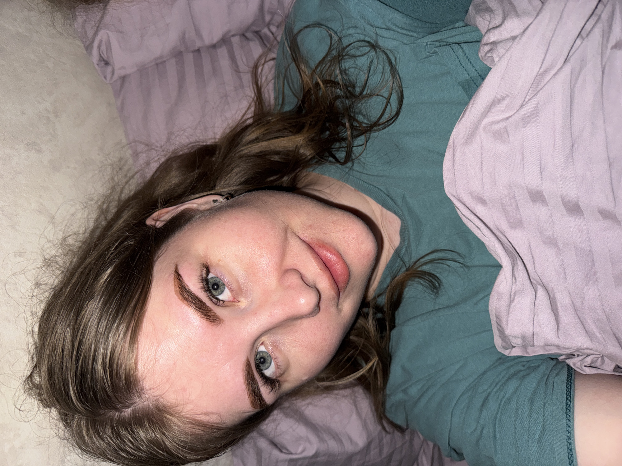 A person with fair skin and light brown hair lies on a bed with light purple sheets and pillow. They wear a dark green t-shirt. The photo is taken from above and slightly to the side of the person's face. A part of the bed and wall are visible in the background. The exact location in Gornyi (Dvurechenskoe S/P), Russia, is indeterminable from the image.