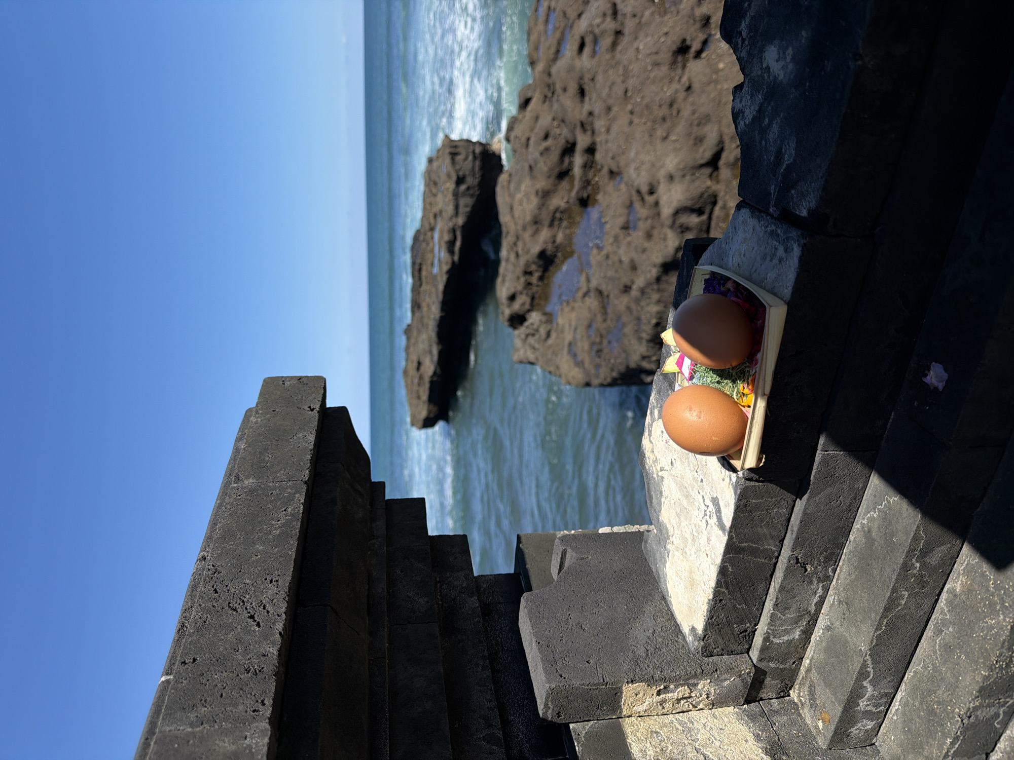 The image shows a Balinese offering, likely a canang sari, placed on a stone structure that appears to be part of a temple or a coastal barrier. The offering contains two brown eggs and possibly other symbolic items like flowers. In the background is the ocean with rocky formations, indicating a coastal environment. The clear blue sky suggests a bright, sunny day. Given the presence of Balinese offerings and coastal landscape, the location is likely North Kuta, Indonesia, an area known for its temples near the coastline.