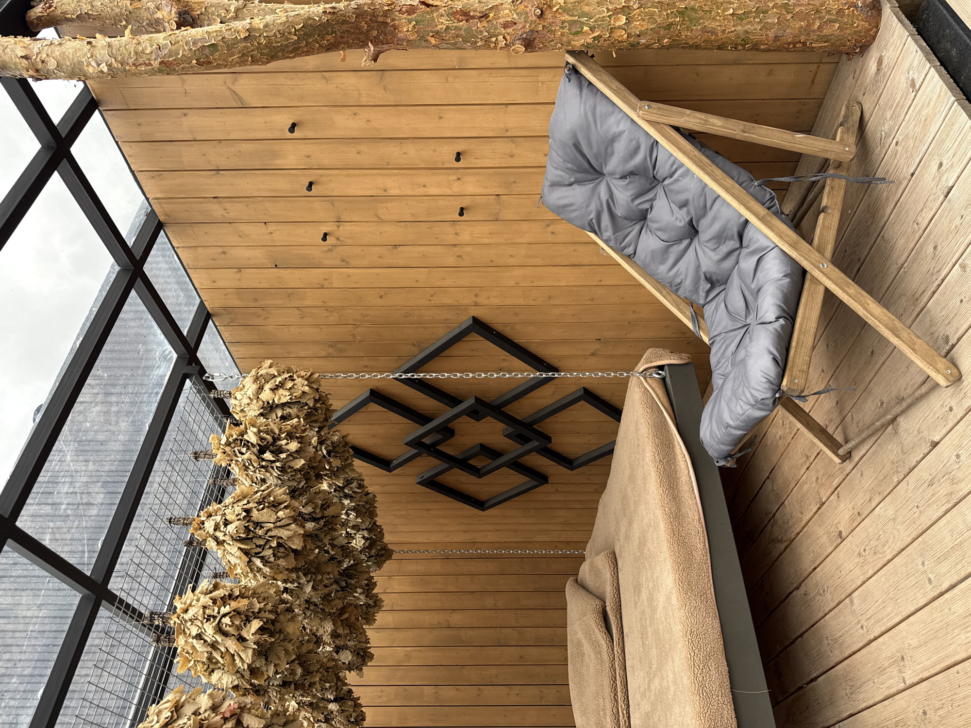 The image shows the interior of a room, possibly a sauna or relaxation space, in Vyakhtelevo, Russia. The walls are constructed of light-colored wood planks. A large window with a black frame is on the left side. Hung on a chain near the window are bundles of dried leaves, potentially birch branches used in sauna rituals. Above, the wooden ceiling has a decorative geometric design made of interconnected black squares. A light-colored wooden chair with a gray cushioned seat is leaning against the right wall. A light tan blanket is draped over what appears to be the top of a bench or cot. A log is above the chair.