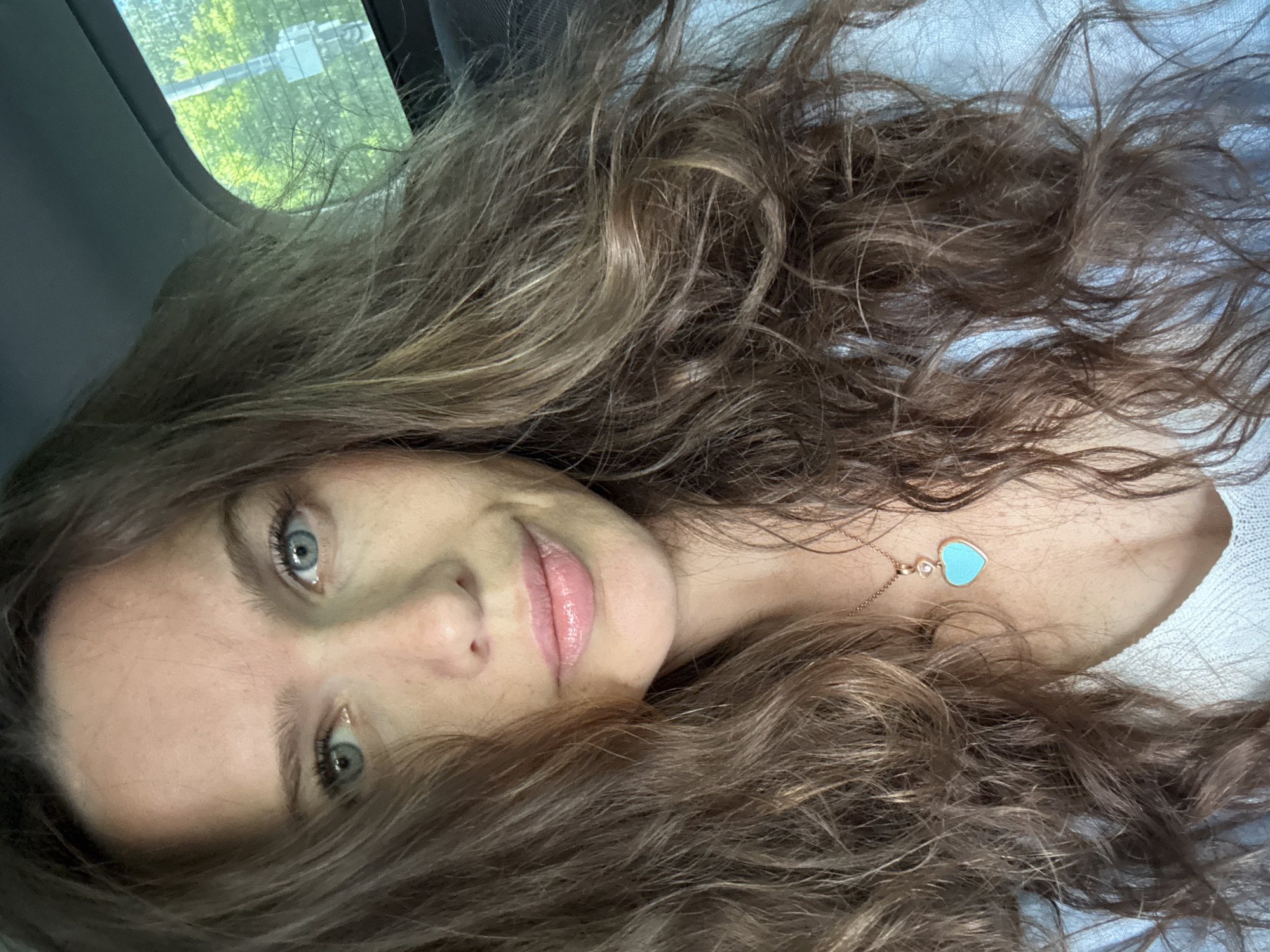 A selfie-style portrait captures a woman with long, wavy, light brown hair, some strands showing lighter highlights. Her head is tilted, and she gazes directly at the camera with clear light blue eyes and a subtle, soft smile on her glossy pink lips. Her hair cascades around her head and shoulders, partially covering a light blue or white textured garment. Around her neck, a delicate rose gold necklace features a small, clear round gem and a prominent light blue heart-shaped pendant.
The setting appears to be inside a vehicle, indicated by the dark gray interior trim visible in the upper left corner. Through a car window, bright green foliage and indistinct blurry shapes can be seen, suggesting the photo was taken during the daytime. The overall impression is one of relaxed composure, with natural light illuminating her features. No visible text is present in the image.