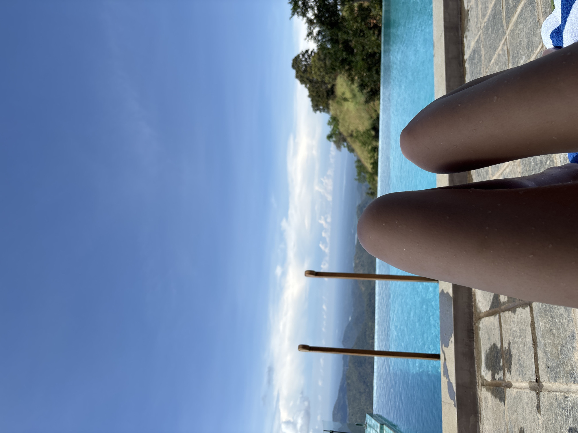 A pair of legs are positioned in the foreground, adjacent to a swimming pool with a clear, blue-green surface. The pool is situated near a ledge with a gray, tiled surface. Beyond the pool, a landscape reveals a mountain range covered in trees and foliage. In the distance, a cloudy sky transitions from a light blue to a deep blue above. The location is Haputhale, Sri Lanka.