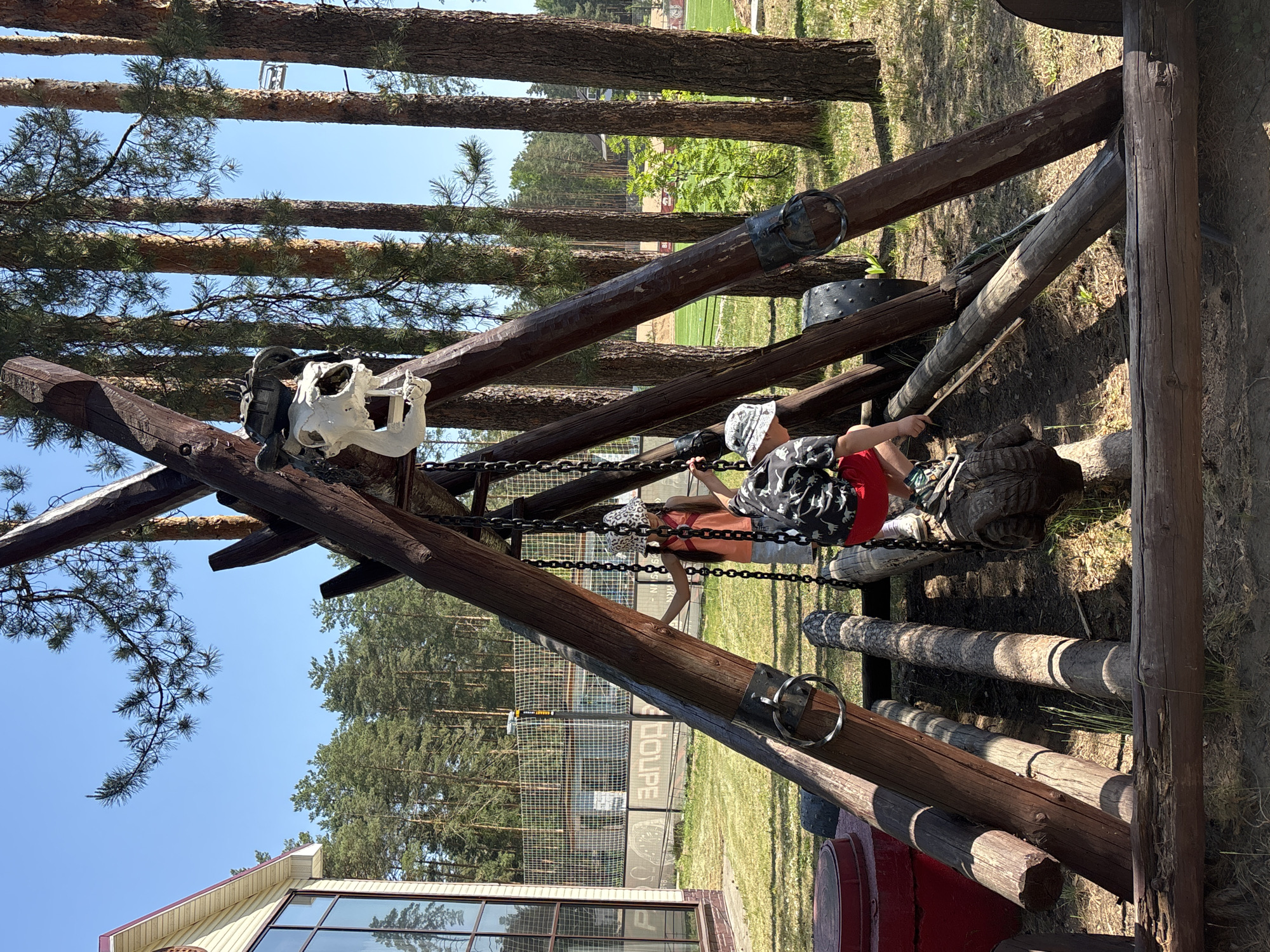 A child is on a playground structure, with trees and a building visible in the background.
**People:** A child, wearing a hat and clothing with a print, is on the playground structure. Another child stands nearby.
**Subjects:** A playground structure made of logs and chains, with a skull decoration on top.
**Objects:** Logs, chains, skull decoration, tires, building with windows, trees, grass.
**Actions:** The child is holding onto the chains of the playground structure.
**Interactions:** The child is interacting with the playground structure.
**Environment:** The scene takes place outdoors, in a grassy area with trees and a building nearby. The sky is blue and visible between the trees. The setting is near Chaykovsky, Russia.