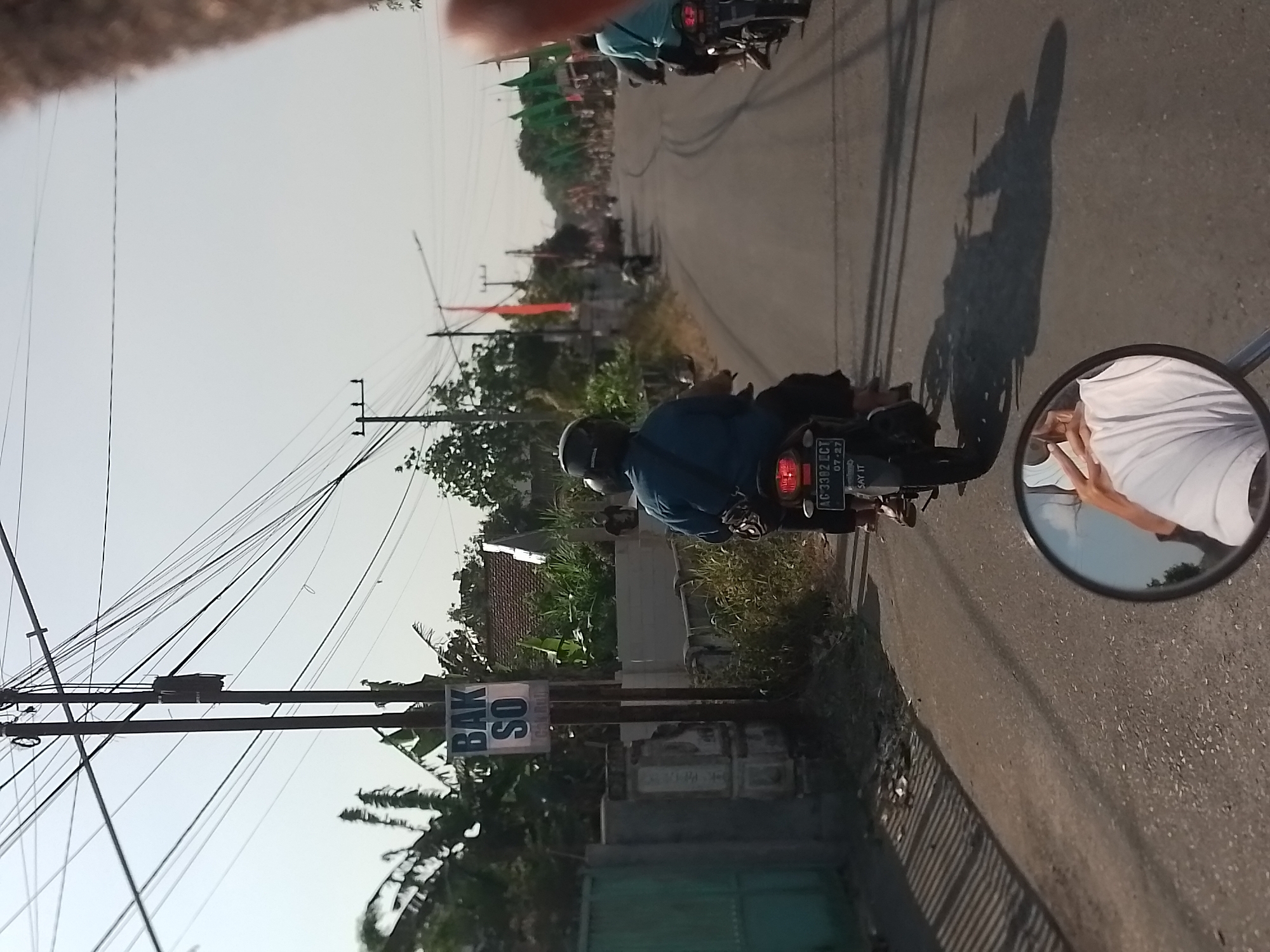 The image captures a street scene in the city of Ngancar, Indonesia, on a clear day. The viewpoint is from the perspective of a person riding a motorcycle, as indicated by the reflection in the rearview mirror showing the rider's white shirt and arm. In the foreground, a motorcycle with a license plate "AG 3392 ECT" and a date "07-27" is visible from the back. The rider is wearing a blue shirt and a dark helmet.
To the left of the street, a utility pole stands tall, adorned with numerous electrical wires and a transformer box. A sign on the pole reads "BAK SO" in large letters. Adjacent to the pole is a green fence and some vegetation.
The street itself is paved and curves to the right. Several other motorcycles are seen on the road ahead. Some are carrying passengers. Flaggers, possibly for a celebration or event, are lined up on the right side of the road, alternating between orange and green colors. Buildings with red tile roofs are visible in the background amidst lush green trees and other foliage. The overall impression is that of a bustling local road in a semi-rural area.