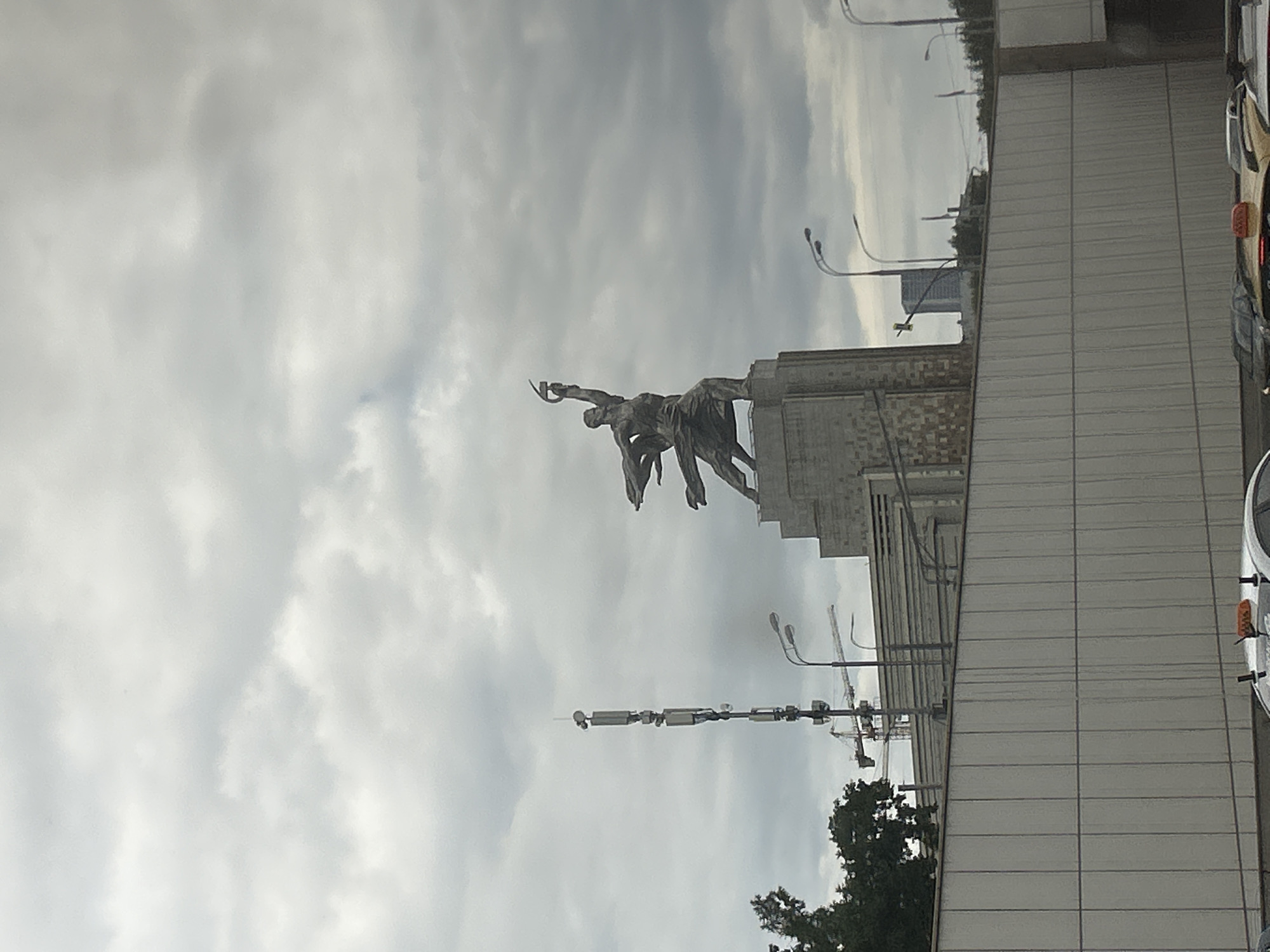 The image depicts a dynamic bronze sculpture of two figures in mid-action, possibly representing labor or sport, with one figure holding a sickle or scythe-like object. The sculpture is mounted atop a large, monumental building with a facade of light-colored, rectangular panels. The building has a classical or socialist realist architectural style, with visible columns or pilasters on the lower section. In the background, a gray, overcast sky dominates the frame, suggesting a cloudy day.
Several streetlights with curved poles are visible, some in the foreground and others further back, indicating an urban setting. Construction cranes can be seen in the distance, hinting at ongoing development or urban renewal in Moscow, Russia. A row of parked cars, including a light-colored vehicle with a visible taillight, lines the side of the building, suggesting a street or parking area. Trees with dark green foliage are also present, providing a contrast to the urban structures. The overall mood is somewhat somber due to the cloudy weather, but the sculpture itself conveys a sense of strength and movement. No visible text is present in the image.