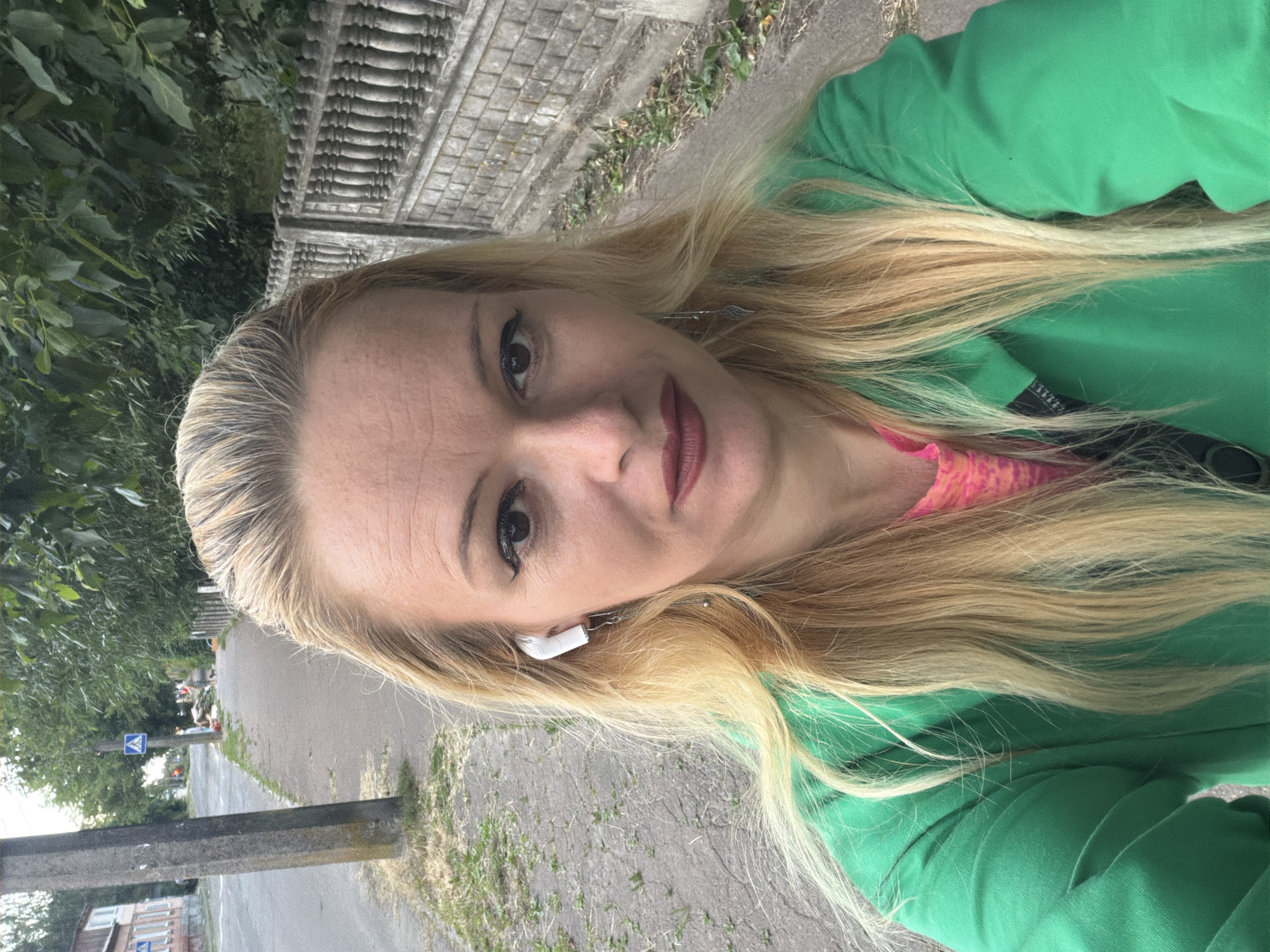 A woman with blonde hair, wearing a green zip-up jacket over a pink shirt, is captured in a selfie. She has on white wireless earbuds and is wearing dangling earrings. The background reveals a sidewalk with a paved surface, bordered by a stone wall with decorative pillars on the right and a row of green trees behind it. To the left, a paved road is visible, with a utility pole and a blue pedestrian crossing sign. In the distance, buildings and more trees can be seen, suggesting an outdoor urban or suburban environment in Chernihiv, Ukraine. The lighting suggests it is daytime.