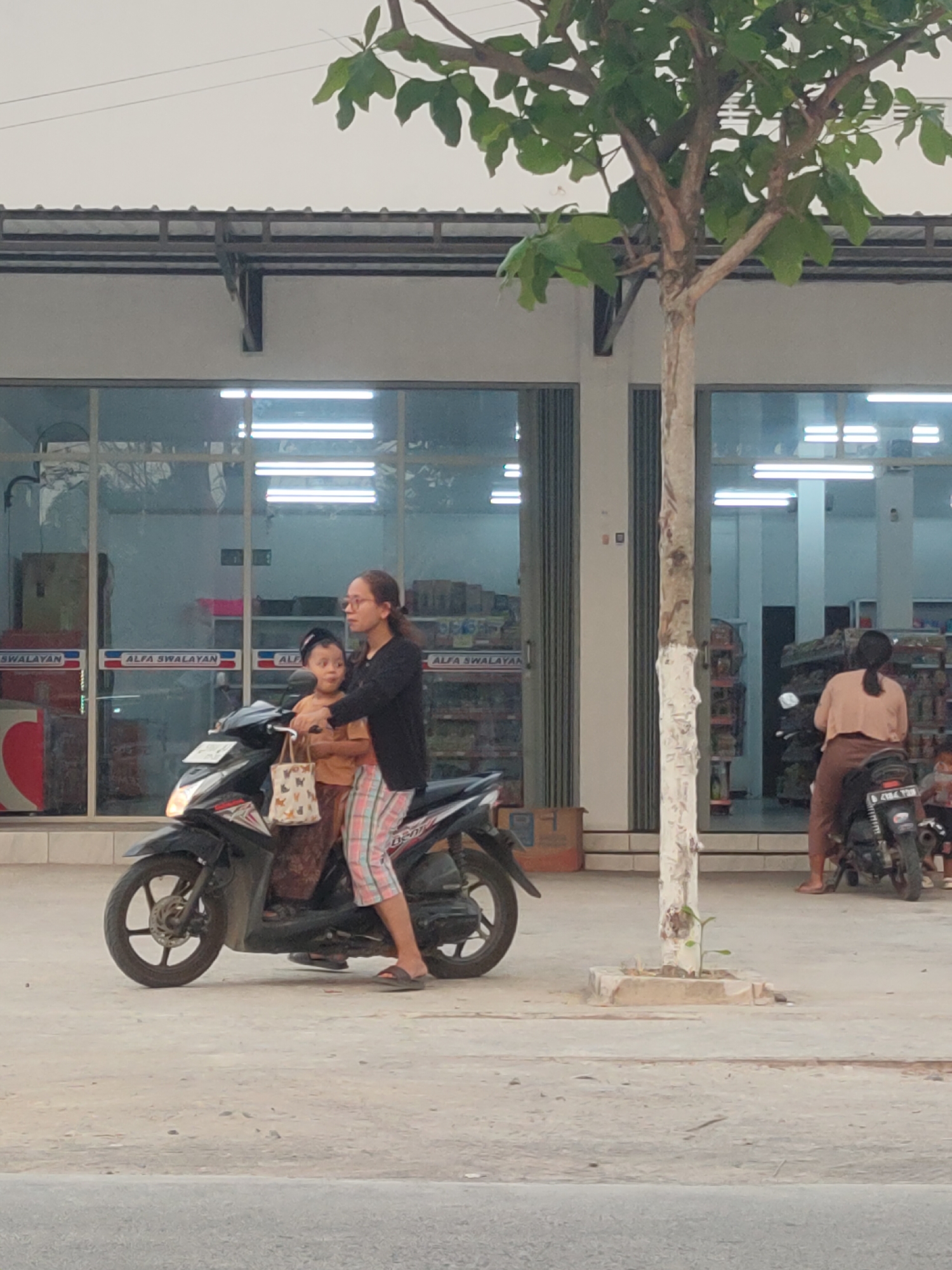 A medium shot captures a paved street in Karanglangu, Indonesia, with a tree in the foreground and a storefront in the background. Two individuals, likely a mother and child, are seated on a black motorcycle parked on the street. The person in the driver's seat, wearing glasses and a black top with checkered pants, appears to be looking forward. A younger individual, wearing an orange top and a brown skirt, is seated behind them, holding onto the driver's shoulders. A white tote bag with a butterfly pattern hangs from the handlebars. The storefront behind them features large glass windows displaying products and fluorescent lighting. Another motorcycle is visible to the right, occupied by a person wearing a pink top and brown pants, facing away from the camera. The ground is a light gray concrete surface, and the sky is overcast.
