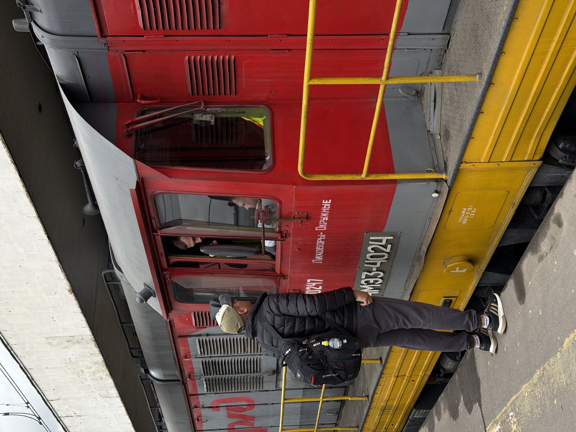 The image depicts a red and grey locomotive parked alongside a concrete platform, likely at a train station or rail yard. A person stands upright next to the train, wearing a black puffer jacket, dark grey pants, and black and white sneakers. A beige baseball cap is on their head, and a black backpack with a water bottle attached is slung over their shoulder.
The locomotive features a vibrant red upper body and a dark grey lower section and roof. A bright yellow safety ladder or railing is affixed to its side. Through the windows of the train's cab, two individuals are partially visible, looking out.
The scene appears to be set during daytime, under what seems to be an overcast sky, as there are no prominent shadows. The ground beneath the platform is asphalt or concrete.
Several Cyrillic inscriptions are visible on the train. On the red body, "0247" and "ЛИХОБОРЫ- ОКРУЖНЫЕ" (LIKHOBORY - OKRUZHNYE) are prominent. A number plate clearly displays "ЧМЭ33-4024" (ChME33-4024), identifying the train's model and serial number. On a yellow section below, text includes "ОБЩИЙ ВЕС T-3/A" (General weight T-3/A) and "123". A faint "2382" can also be seen on the lower grey part of the train. The setting is a railway environment, consistent with Moscow, Russia.