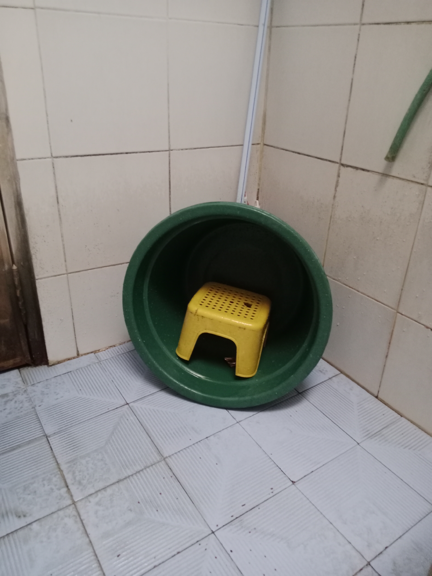 A yellow plastic stool is placed inside a large, green plastic basin. The basin is positioned on a tiled floor in what appears to be a bathroom or laundry area. The walls are covered in white square tiles with dark grout lines. A green hose is visible attached to the wall in the upper right corner. A doorframe with a dark wooden door is on the left side of the image. The floor tiles have a patterned texture. The scene is located in the city of Kediri, Indonesia.