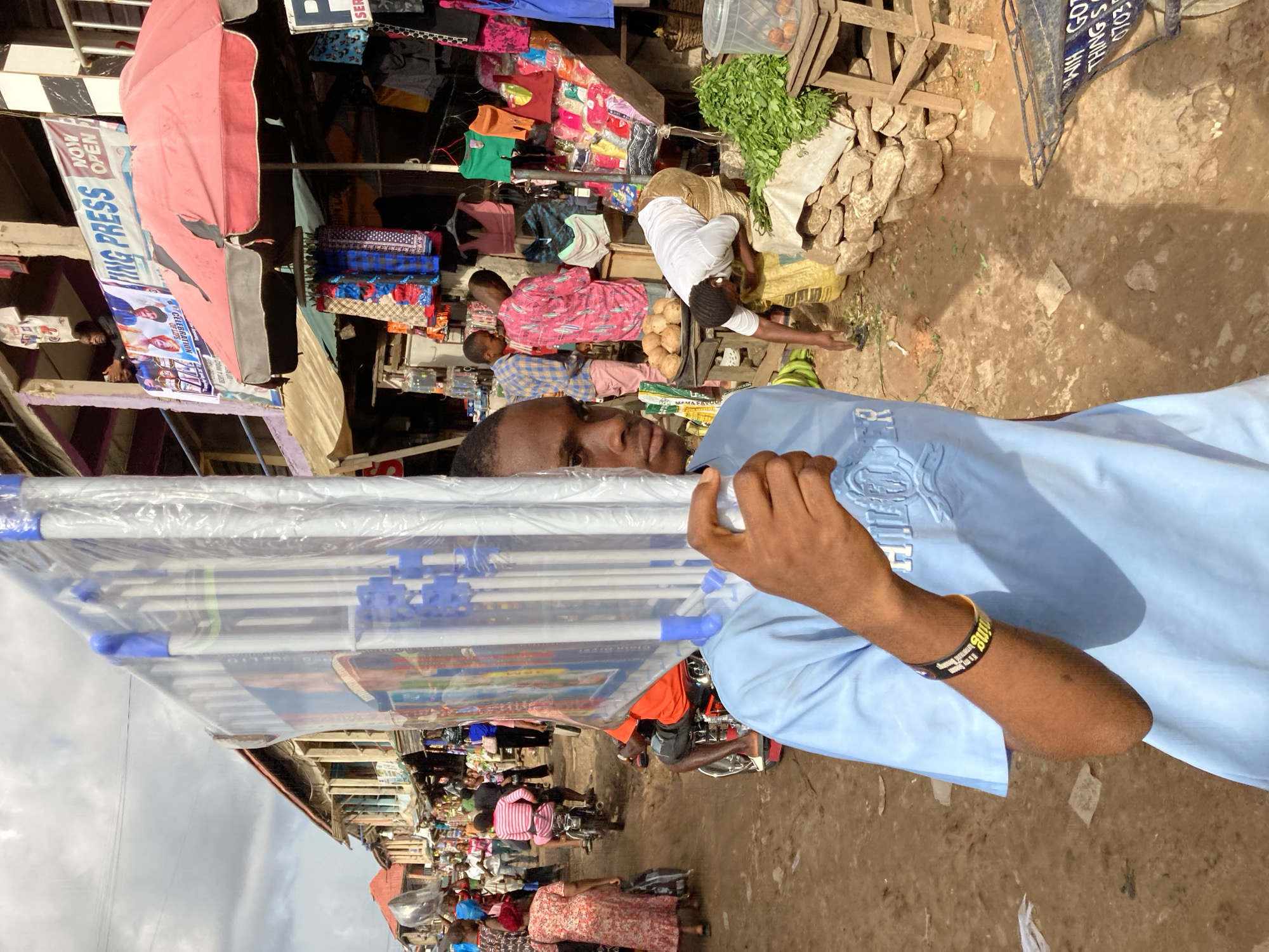 The image shows a bustling marketplace in Ura Nta Umuarandu, Nigeria. A man in a light blue shirt and a black and gold bracelet is in the foreground, holding a package of what appears to be collapsible clothes drying racks wrapped in plastic. Behind him, the marketplace is crowded with people. Several vendors are visible, including one selling leafy greens and possibly other produce. There are clothing items hanging from stalls or shop awnings. The ground appears to be dirt or a worn pathway. In the background, there are various buildings and structures, including a sign for "ING PRESS" that also states "NOW OPEN."