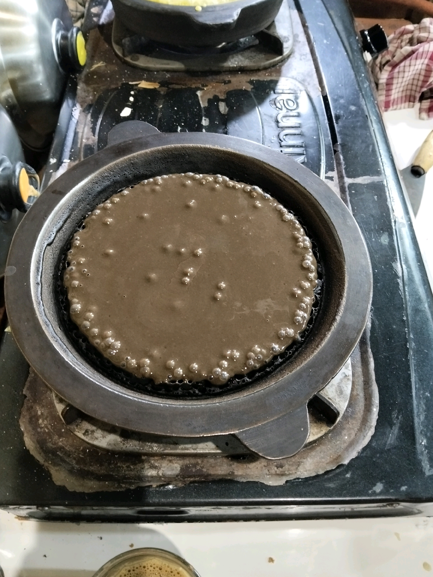 A overhead shot shows a circular pan on a black gas stove. The pan is filled with a dark brown, liquid batter, which is beginning to bubble around the edges. The batter has small, evenly distributed bubbles throughout. The stove appears to be used, with some discoloration and debris around the burner. To the right of the stove, a red and white checkered cloth is visible, along with a wooden-handled object. In the lower part of the image, a glass containing a frothy, light brown beverage is present. The overall scene suggests food preparation in a domestic setting. The exact location of Arang Limbung, Indonesia is not depicted in the image.