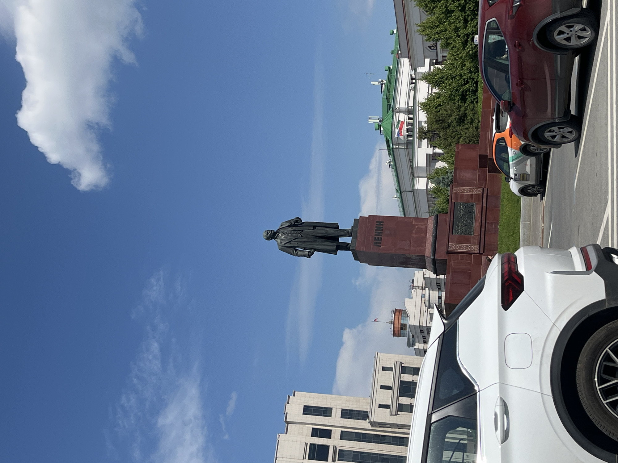 The image shows a statue of Lenin in Kazan, Russia. The statue is located in a prominent square, with several buildings visible in the background, including the National Library of Tatarstan. The sky is blue with some clouds. There are cars in the foreground, suggesting that this is a public space. The overall impression is one of a grand, historical monument in a modern urban setting.
