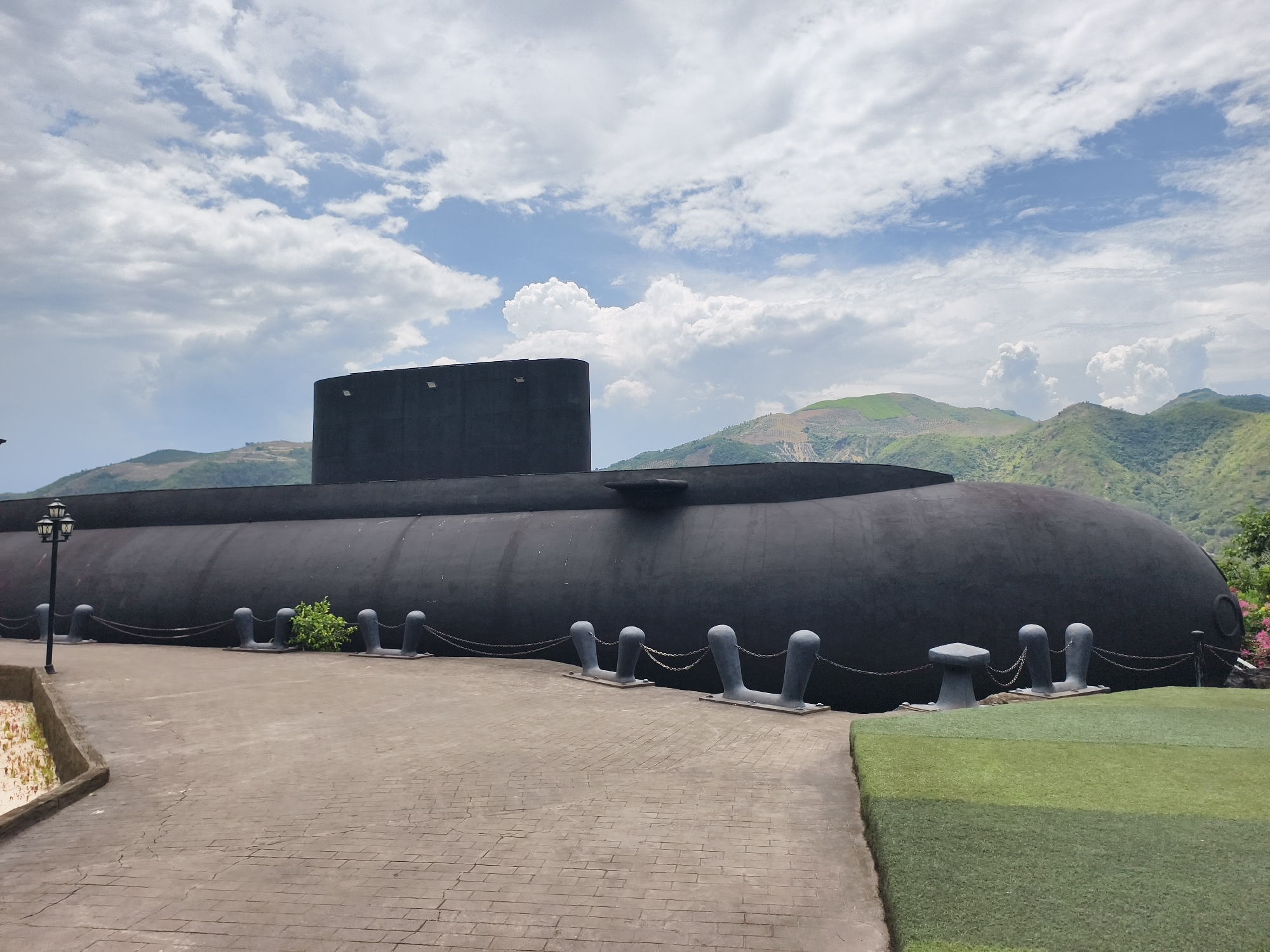 The image shows a black submarine displayed on land in Tay Nha Trang Ward, Vietnam. The submarine has a prominent conning tower. It is positioned on a paved area next to a section of artificial turf. Mooring bollards are arranged along the side of the submarine, connected by chains. A small green bush grows between two of the bollards. In the background, mountains are visible under a sky filled with white clouds. A street lamp is visible to the left of the submarine.
