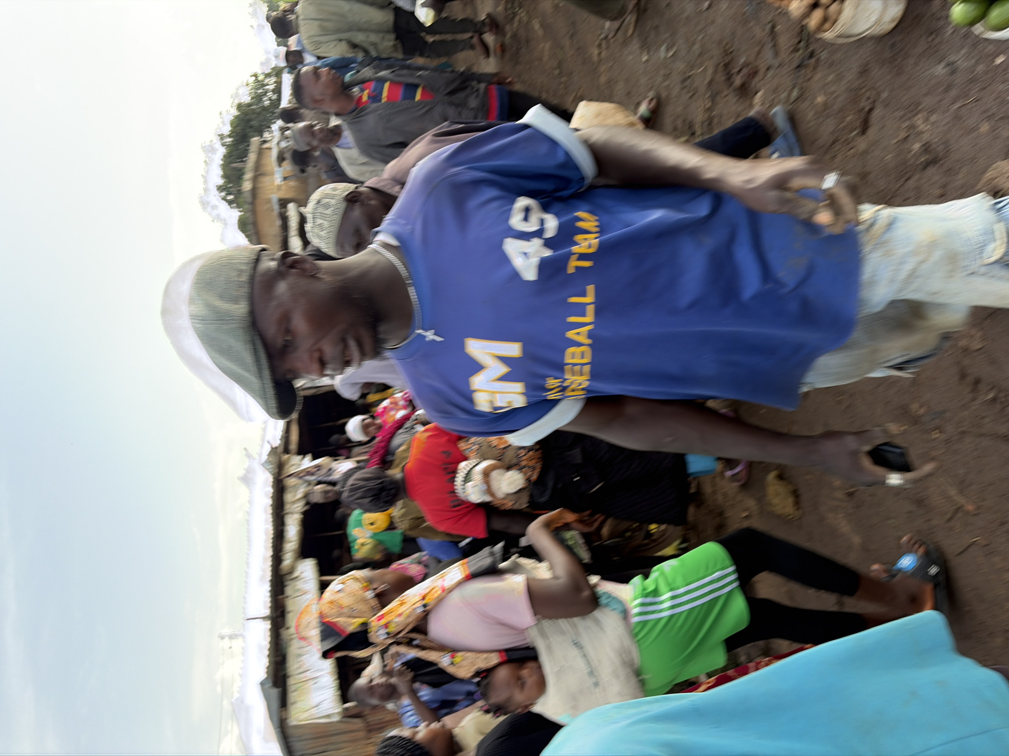 The image depicts a bustling outdoor market scene in Gura Top, Nigeria, with a focus on the foreground. A man wearing a green and white flat cap, a silver chain with a cross pendant, and a blue t-shirt with yellow lettering that reads "EM BASEBALL TEAM 49" is looking to his right. His t-shirt also has a small patch on the left sleeve. He is wearing light-colored, possibly denim, shorts that appear dirty.
To his left, a woman in a pink shirt and a vibrant, patterned headscarf is carrying a baby on her back in a light-colored wrap. She is wearing green shorts with white stripes and black leggings. Another woman with a red shirt and a patterned headscarf is visible behind her, also carrying a baby.
The background is filled with a crowd of people, some of whom are obscured. Structures that appear to be market stalls or makeshift shops made of corrugated metal and wood are visible. The ground is a mix of dirt and some scattered debris. In the lower right corner, a white bucket filled with what appear to be fruits or vegetables is visible, and some green fruits are on the ground. The overall impression is one of lively commerce and community activity.