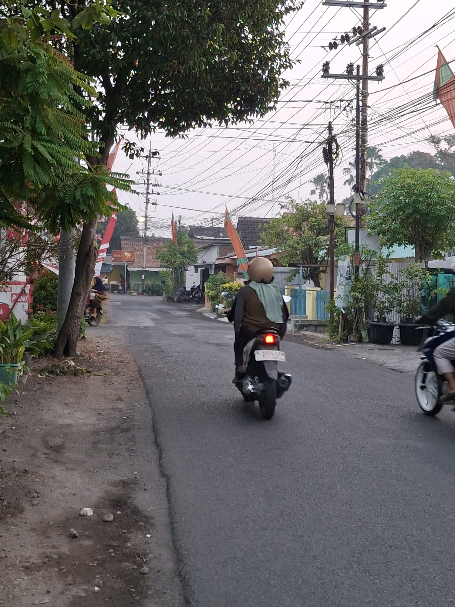 The image depicts a street scene in Lumajang, Indonesia, likely in a residential area. The road is paved with asphalt and appears to be somewhat narrow. On the right side of the road, there is a motorcycle with a rider wearing a brown jacket and a light green scarf, and a helmet. The motorcycle's taillight is illuminated red. Further down the road, another motorcycle is partially visible on the right, with its front wheel and rider's legs showing. On the left side of the road, there is a dirt and gravel shoulder with some vegetation and a tree. Electrical poles with numerous wires are prominent features of the environment, stretching across the upper portion of the image. Buildings with tiled roofs are visible in the background, suggesting a built-up area. Several flags, possibly decorative or celebratory, are also present, attached to poles along the street. The overall lighting suggests it is either early morning or late afternoon.