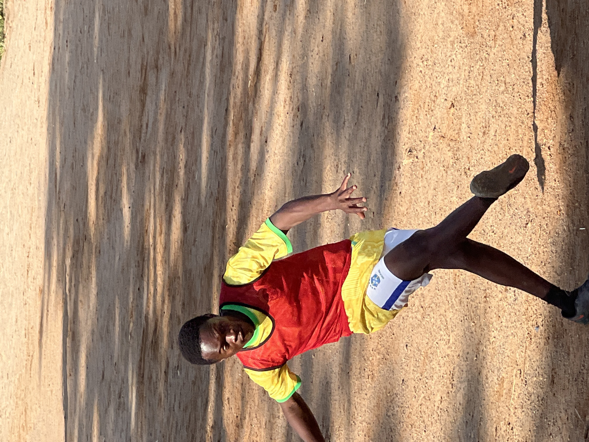 A young Black male is shown playing soccer on a dirt field in Jos, Nigeria. He is wearing a yellow soccer jersey with green trim, a red training bib, and white shorts with blue trim and a Brazilian flag emblem. His left leg is bent and positioned behind him as if he is about to kick a ball, while his right foot is extended forward. His hands are outstretched, with his left hand extended further forward than his right, likely for balance. The ground is composed of reddish-brown dirt with scattered shadows cast by unseen objects, suggesting an outdoor, possibly sunny environment. The background is blurred, with the focus on the individual and his action.