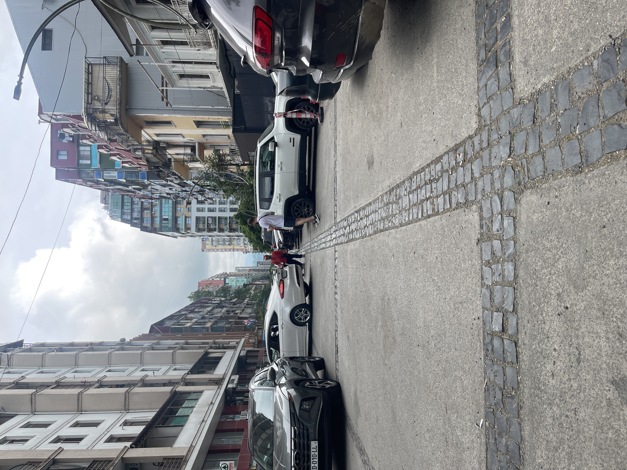 This media file, taken in Batumi, Georgia, depicts a street scene with multiple vehicles and buildings. The image is oriented vertically. In the foreground, a concrete sidewalk is marked with a cross pattern formed by dark gray cobblestones. To the left of the sidewalk, several cars are parked. A white SUV is prominently featured, with red and white tape wrapped around its front tires, suggesting it might be cordoned off or awaiting inspection. A person in a red jacket and shorts is standing near the white SUV, appearing to interact with it or the surrounding area. Further down the street, more vehicles are visible, including a dark gray SUV in the foreground.
The background is filled with multi-story buildings, characteristic of an urban environment. These buildings vary in architectural style and color, with some featuring balconies and numerous windows. The sky is overcast, indicating a cloudy day. The overall scene suggests a typical day in a city, with a focus on the street and parked vehicles. No explicit time of day is indicated, but the lighting suggests it is daytime. The presence of the parked cars and pedestrian activity points to an active urban setting. Some text is visible on a license plate of the dark gray SUV: "TT-010-LL".