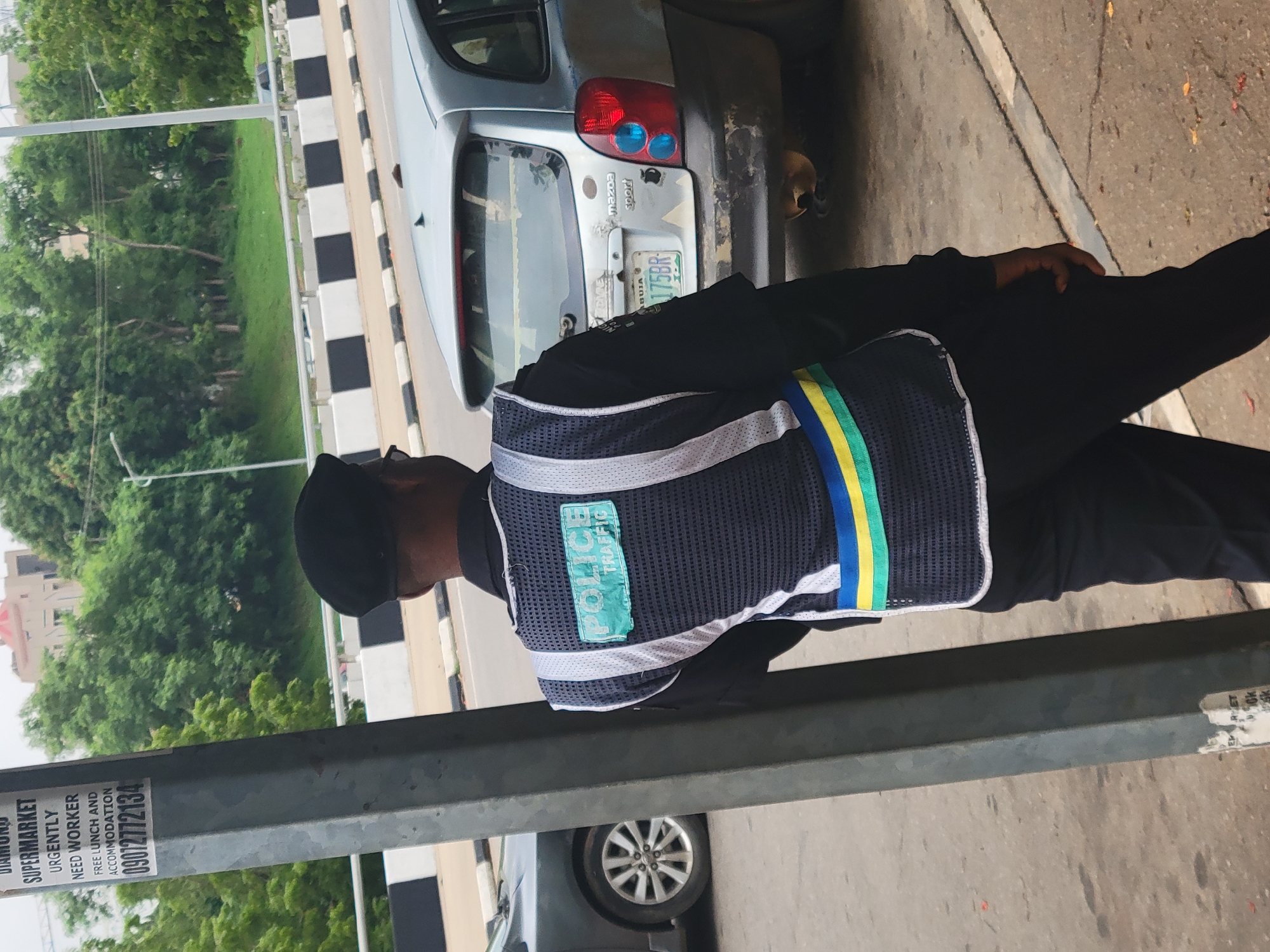 A police officer stands near a utility pole in what appears to be an urban setting in Abuja, Nigeria. The officer is wearing a dark uniform with a reflective vest that has "POLICE TRAFFIC" printed on it in green lettering. The vest also features reflective white stripes and a horizontal band of blue, yellow, and green. The officer is looking away from the camera, towards the right side of the frame.
In the background, a silver-colored hatchback vehicle is parked on the side of the road. The vehicle's license plate is visible and appears to read "ABUJA 175BR". Beyond the road, there is a grassy embankment with trees and some buildings in the distance. The ground in the foreground is paved, with a visible road marking and some fallen leaves. A portion of a large, grey utility pole is prominently positioned in the left of the frame, with a flyer attached to it that advertises a "DIAMOND SUPERMARKET" seeking workers.