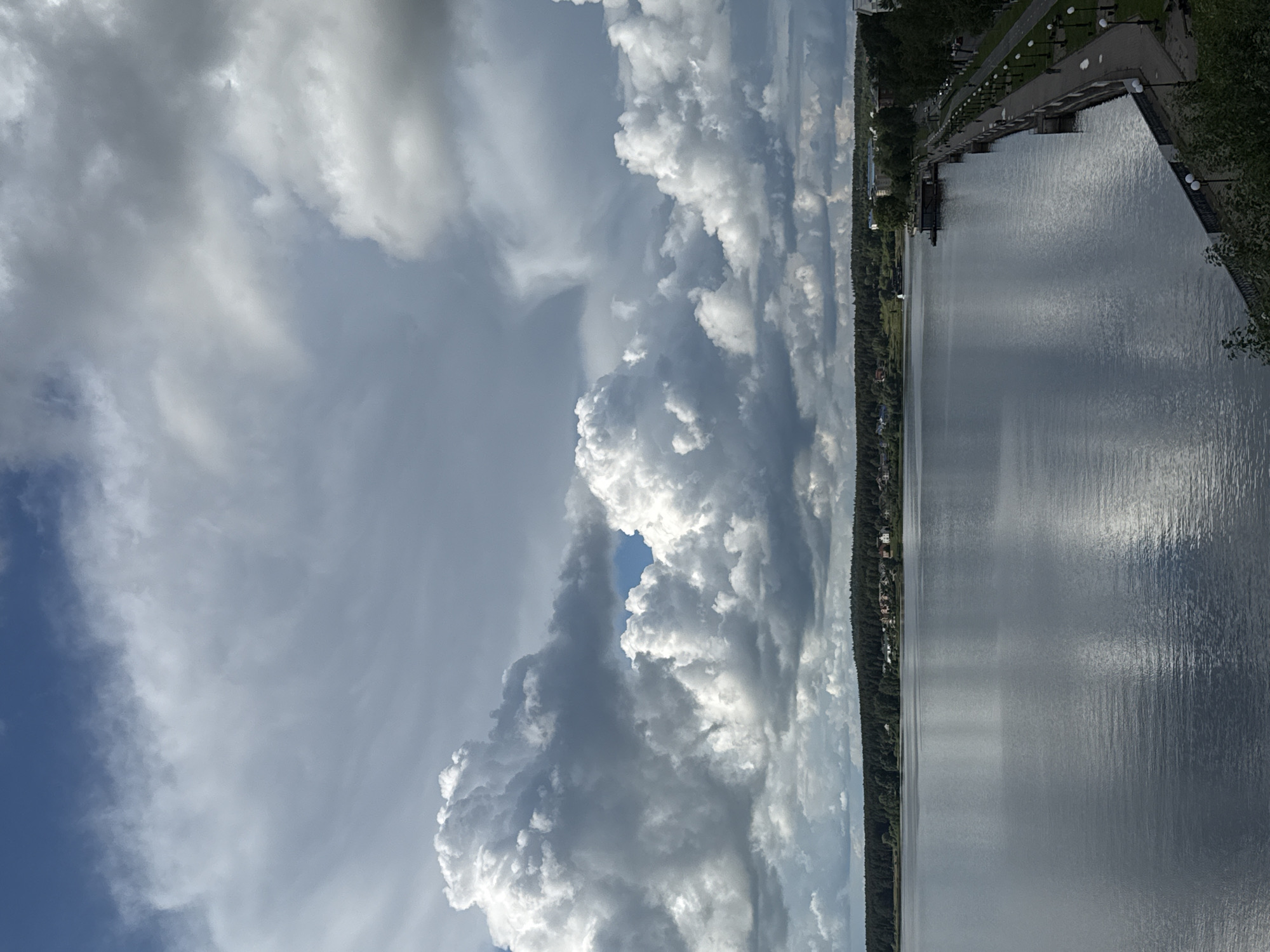 The image, when viewed in landscape orientation, captures a scenic waterscape under a dramatic sky, located in Votkinsk, Russia. The perspective appears to be from an elevated position overlooking a wide body of water, possibly the Votkinsk Reservoir.
The upper portion of the frame is dominated by a dynamic sky filled with large, bright white cumulus clouds. These clouds are voluminous and textured, displaying varying shades of grey where light is less direct and brilliant white where illuminated by the sun. Patches of clear blue sky are visible between the impressive cloud formations, particularly towards the upper left.
The middle and lower-middle sections of the image feature a wide expanse of water, appearing calm with gentle ripples. The surface of the water reflects the sky and clouds, showing distinct lighter and darker streaks. The water generally has a silvery-grey hue under the partly cloudy conditions.
On the right side of the water, a developed embankment or promenade is visible, running parallel to the water's edge. This area includes a paved pathway lined with numerous lampposts or light fixtures, suggesting an urban park or waterfront area. Trees and lush greenery border this path. On the opposite, more distant shore, a long stretch of dense forest defines the horizon line, with scattered residential buildings or structures faintly discernible within or at the edge of the tree line.
No people or vehicles are visible in the scene. The time of day appears to be daytime, likely late morning or afternoon, given the bright, well-defined clouds and reflections. The weather is partly cloudy, creating an interesting play of light and shadow. The overall scene conveys a sense of tranquility and the grandeur of nature, with the dramatic cloud formations being a notable focal point. There is no visible text.