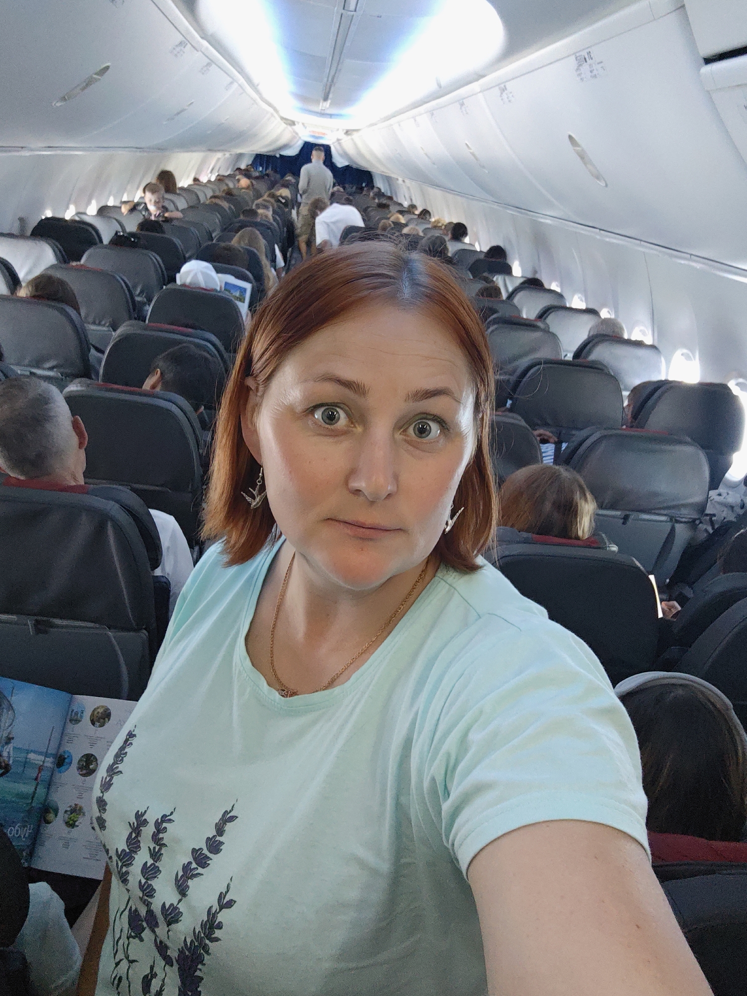 The image shows a woman with reddish-brown hair taking a selfie in the aisle of an airplane. She is wearing a light blue t-shirt with a floral pattern and a gold necklace. Her expression is one of surprise or mild alarm, with her eyes wide open and her mouth slightly ajar. She is facing the camera, and the background shows rows of airplane seats filled with passengers. The overhead compartments and lighting of the airplane cabin are visible, suggesting it is a typical commercial flight. The overall scene is indoor, within the confines of an aircraft.
