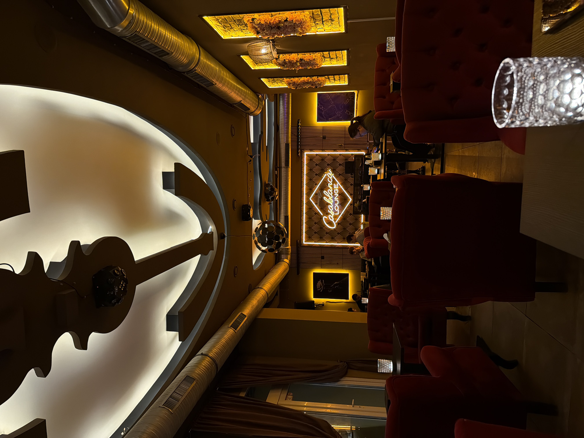 The image captures the interior of the Casablanca Lounge & Bar, located at Budapester Str. 38-40, 10787 Berlin, Germany. The scene depicts a dimly lit lounge environment illuminated by warm, amber-toned artificial light.
The ceiling features elaborate, undulating wooden architectural elements with recessed strip lighting, a central spherical metallic light fixture, and a smaller, black multi-faceted disco-ball-like projector. Exposed corrugated metal air ducts run horizontally along the upper sections of the walls. The walls are painted a muted olive-brown.
Seating predominantly consists of red velvet high-backed armchairs and matching sofas, arranged around small, dark, square tables throughout the lounge. Each visible table holds a clear, textured glass votive with a lit candle.
Approximately five to seven individuals are visible. On the right, a woman with dark hair tied back is seated at a table, looking down at a device. Towards the rear-left, two men are seated at a table, one in a light-colored hoodie, both appearing engaged with personal devices or in conversation. Other patrons are partially obscured in the background. No direct interactions or specific activities are clearly discernible among them.
Prominent decorative elements include a large neon sign centrally positioned on the back wall, outlining a diamond shape and spelling "Casablanca LOUNGE" in script. To its left, a framed dark art piece features a neon-outlined rose. To the right, multiple vertical rectangular panels are affixed to the wall, featuring backlighting that illuminates faux brick textures and artificial floral arrangements. A large window or glass door with heavy brown drapes is visible on the far left. The floor is tiled with light-colored, square tiles.